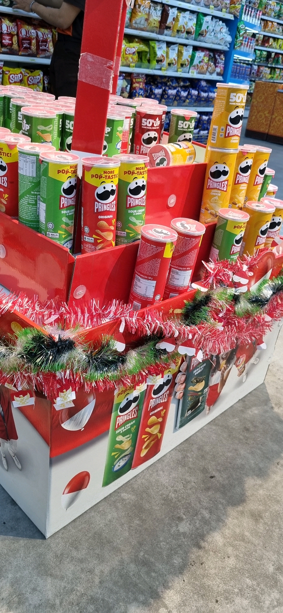An exterior shot shows a store display of Pringles potato chip cans, arranged in a red cardboard box. The cans are stacked and positioned upright, featuring various flavors such as Original, Sour Cream & Onion, and Cheesy Cheese, with the Pringles mascot visible on each can. The display is decorated with red and green tinsel, and small Santa hats are attached to the tinsel, indicating a festive or holiday theme. In the background, shelves are stocked with various snack food items, and a person wearing a black shirt and orange vest is partially visible, suggesting a retail environment. The ground is concrete, with shadows cast from the display and surrounding structures. The location is identified as Denpasar, Indonesia, possibly a supermarket or convenience store.
