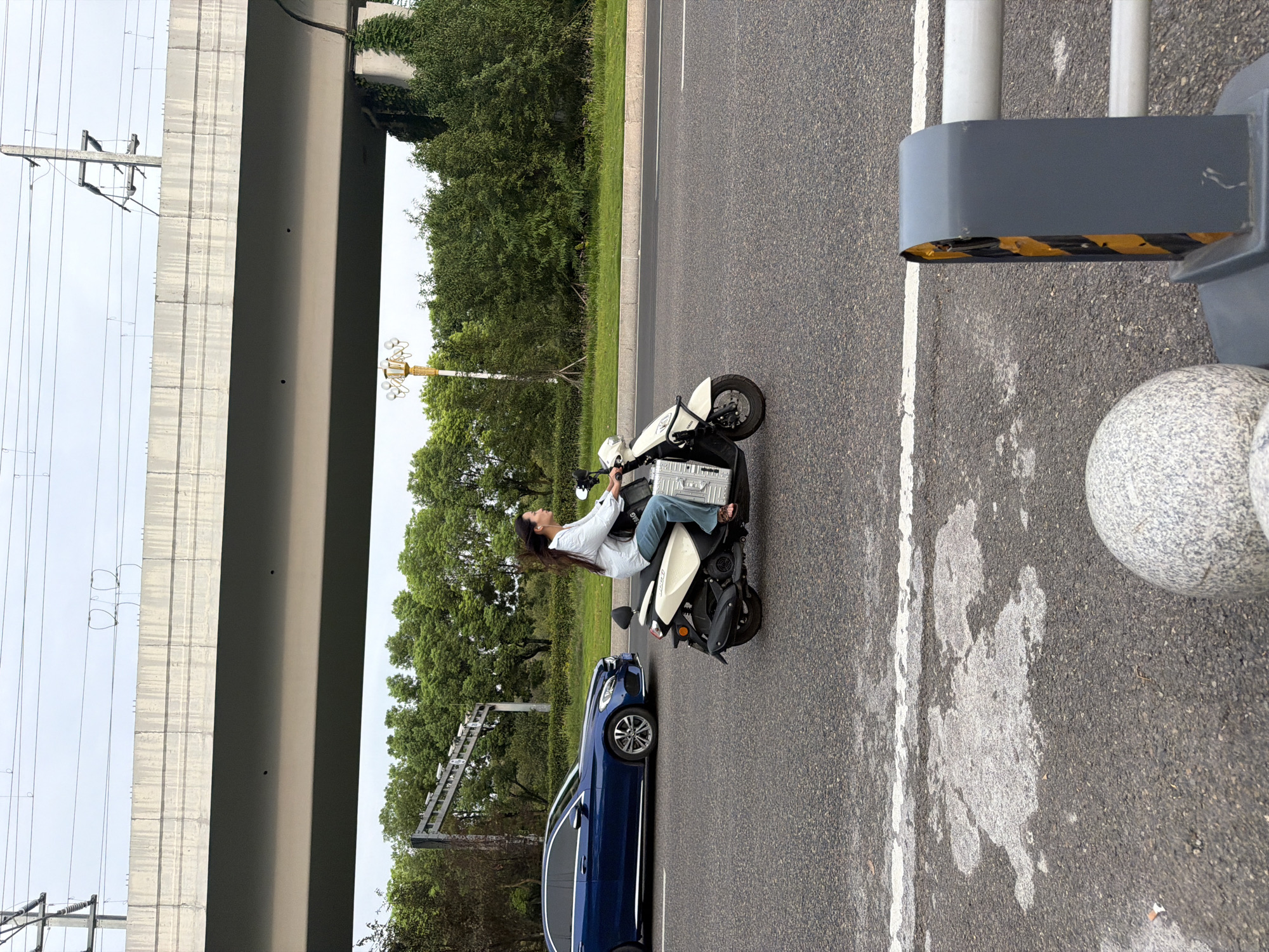 A woman with long dark hair pulled back in a ponytail rides a cream-colored scooter on a paved road in Jiangjin, China. She is wearing a light blue long-sleeved shirt and blue jeans. The scooter has a silver luggage case attached to the back. To the left of the scooter, a dark blue Mercedes sedan is driving in the same direction. In the background, there is a large concrete overpass with electrical wires and poles overhead. Lush green trees line the side of the road. In the foreground, a section of a guardrail with black and yellow hazard stripes is visible, along with a spherical stone object. The sky is overcast.