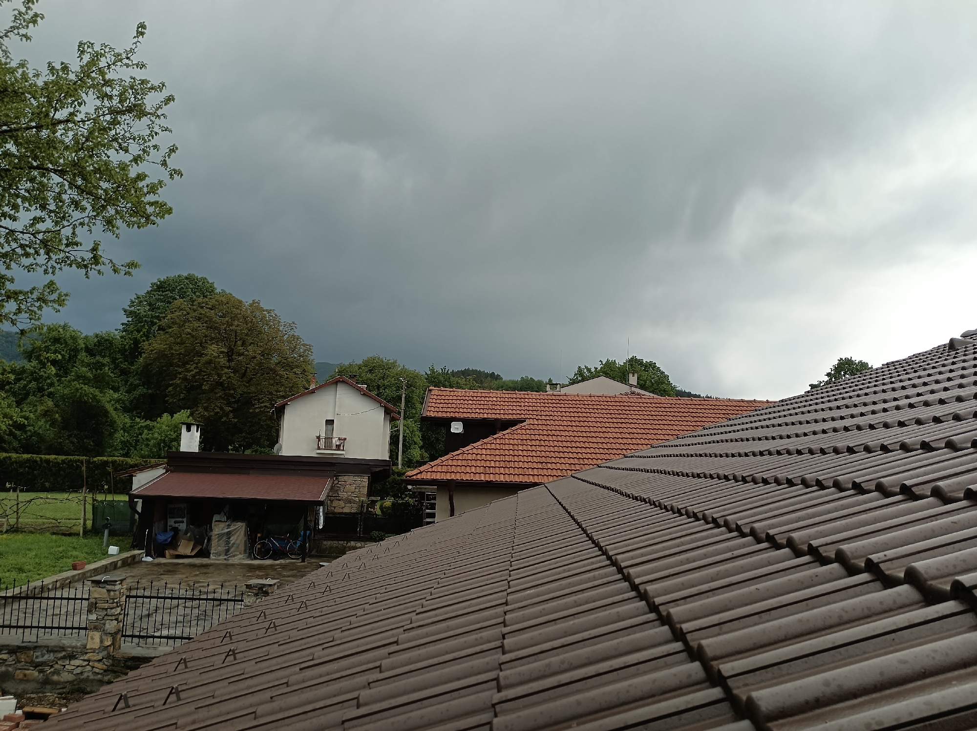 The image depicts an outdoor scene, likely taken on a rainy or overcast day due to the dark and cloudy sky. In the foreground, a brown tile roof dominates the view. In the mid-ground, there are several houses with terracotta tile roofs and white walls, surrounded by trees and greenery. The location is Mihaylovtsi, Bulgaria. Other objects include a stone and iron fence and a bicycle. There are no people visible in the image. The environment suggests a residential area in a rural or suburban setting.