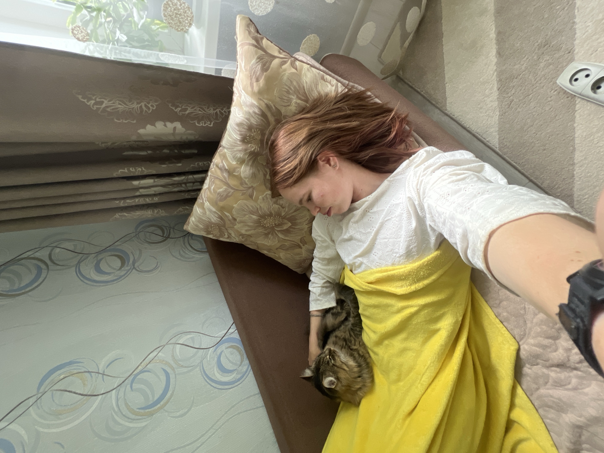 The image presents an intimate indoor scene in Chaikovsky, Russia, centered on a young woman and a cat resting peacefully on a sofa. The woman, with reddish-brown hair, is lying on her left side, seemingly asleep or deeply relaxed, with her head propped on a large decorative pillow adorned with a gold and brown floral pattern. She is wearing a white long-sleeved top with textured or embroidered detailing. A bright yellow blanket covers her lower body and legs, under which a long-haired tabby cat is snuggled closely to her, appearing to be asleep or dozing.
Natural light streams in from a window in the background, suggesting it is daytime. The window is partially obscured by light brown Roman blinds featuring a subtle, ornate pattern. Beyond the blinds, some green foliage can be seen. The room’s decor includes a light blue wallpaper with abstract grey and blue swirling patterns on the wall adjacent to the sofa, and a cream-colored rug with vertical stripes is visible on the floor. A white Type F power outlet is mounted on a wall in the background, consistent with the specified Russian location. The woman is wearing a dark digital watch on her right wrist. The overall ambiance of the scene is one of tranquility and comfort.
