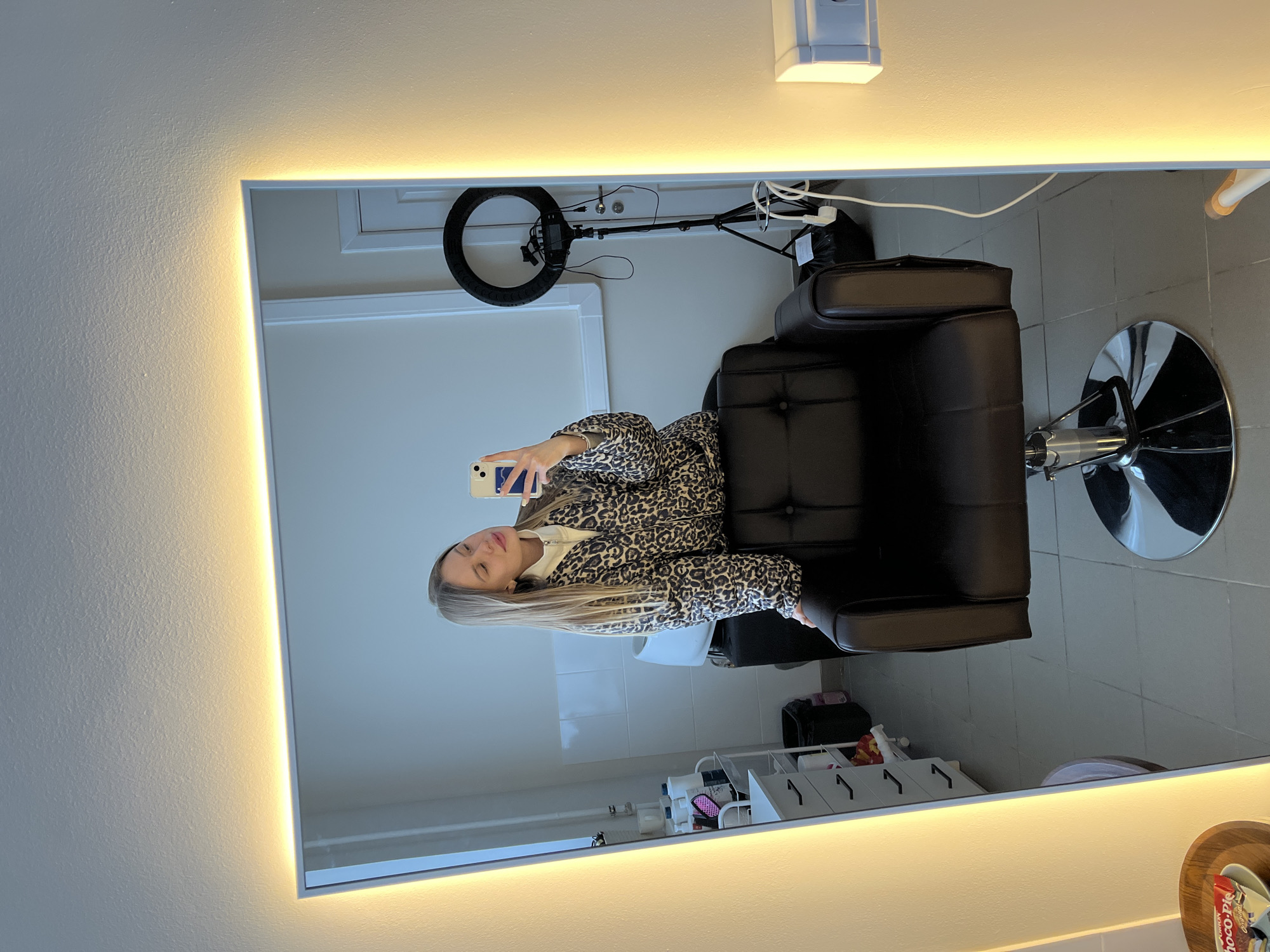 A mirror reflects the interior of what appears to be a salon or similar beauty-related establishment. A woman is seen taking a selfie in the mirror while seated in a black salon chair. The chair is positioned in the center of the reflected image. The salon setting includes a white wall, light fixtures, a ring light, and various salon supplies arranged on shelves. Based on previous information, the location of this scene is Chaikovsky, Russia.
