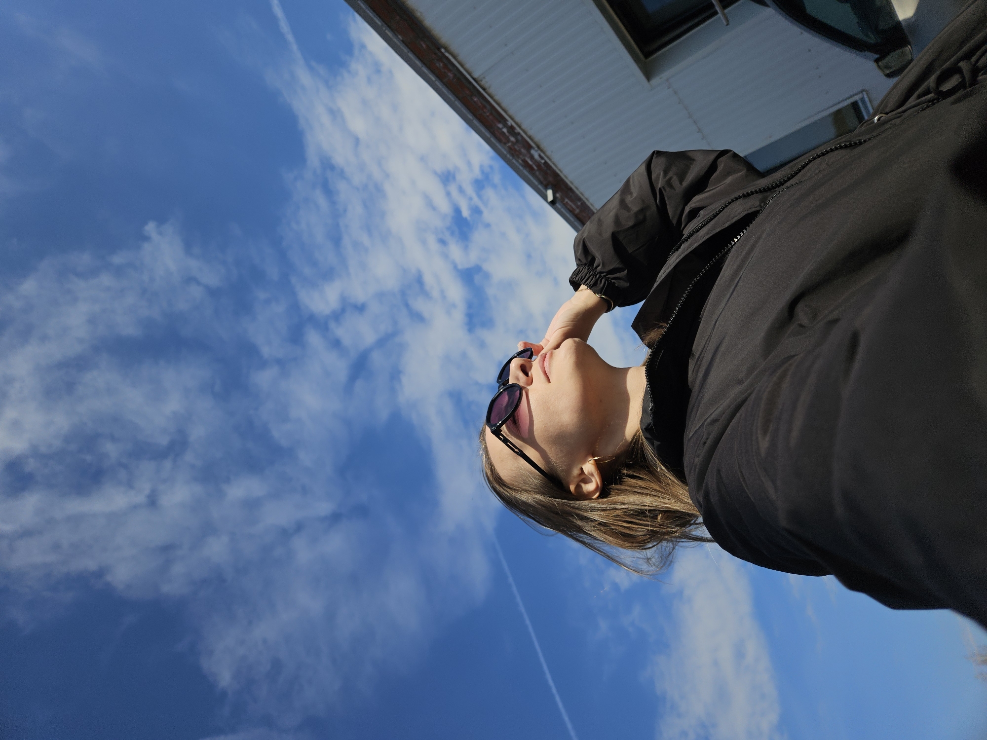 An upward-angle, tilted photograph captures a person against a bright blue sky with scattered white clouds. The subject, likely female, is seen from the chest up, positioned on the right side of the frame. She wears a black puffer jacket and dark sunglasses with purple-tinted lenses. Her right hand is raised to her face, touching the side of her head or sunglasses. Her light brown hair flows back from her face. She appears to be looking upwards, possibly at the sky or sun.
In the background, a clear, sunny daytime sky dominates the scene, featuring wispy white clouds and a faint, thin contrail from an aircraft extending diagonally across the lower left quadrant. A muted building structure, possibly a roofline or upper portion of a house, is visible along the top edge of the frame, tilted significantly due to the camera angle. The building has a light-colored soffit or underside and a darker, possibly brick or wooden, upper section. No text is visible in the image. The setting suggests an outdoor location, likely in Chaikovsky, Russia, enjoying a clear weather day.