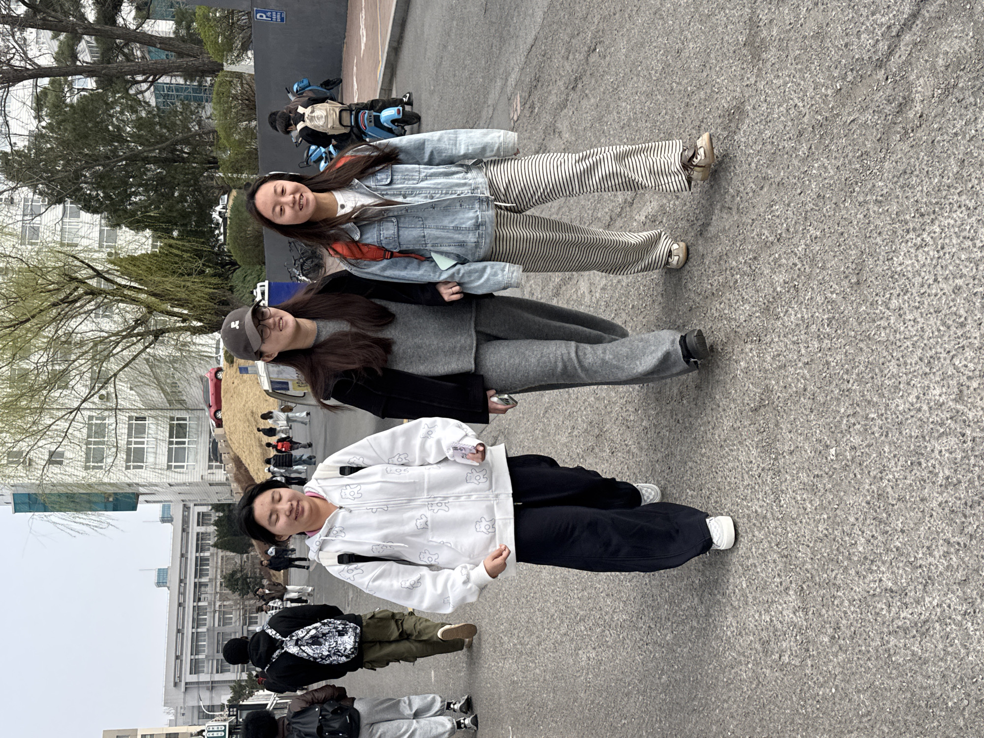 Three women stand together on a paved surface in Zhongtun, China. The woman on the left is wearing a white hoodie, black pants, and white sneakers. The woman in the middle is wearing a gray top and pants with a black jacket and a baseball cap. The woman on the right is wearing a denim jacket and striped pants with tan sneakers. There are buildings and trees in the background. Other people are visible in the background.