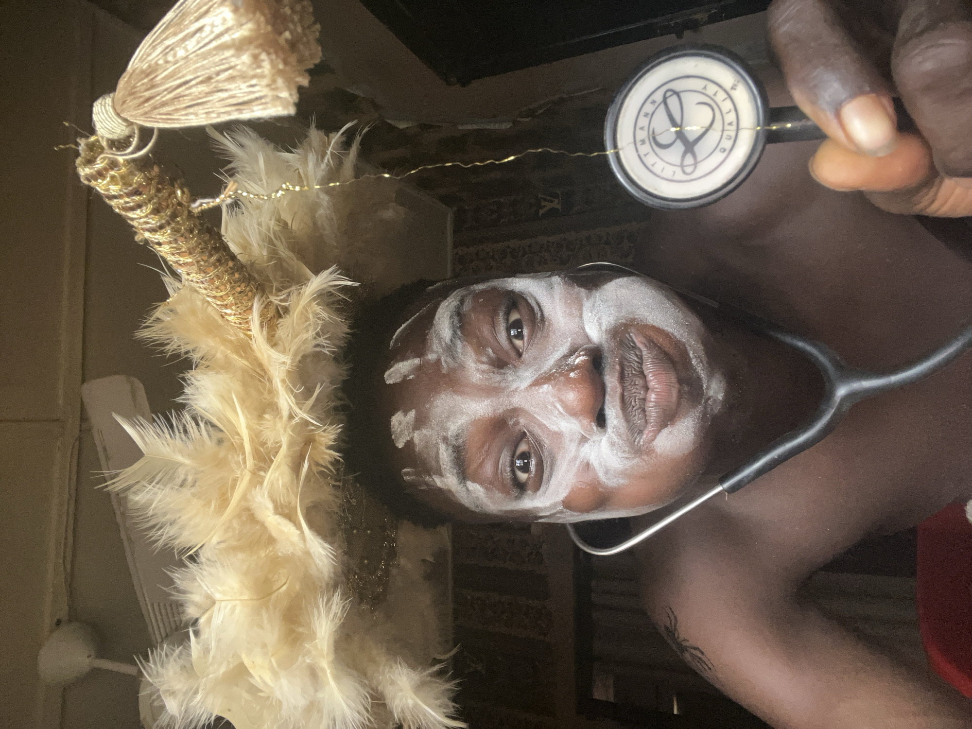 An upright portrait captures a person with dark skin, looking directly at the camera with a neutral and focused expression. Their face is decorated with white paint, applied in distinct patterns across the forehead, cheeks, and around the mouth, suggesting a ceremonial or artistic context.
To the left of their head, an elaborate decorative element is prominent, featuring abundant light-colored (cream or off-white) feathers. This decoration also includes a golden, textured cylindrical piece that appears to be covered in sequins or glitter, topped with a golden tassel. Fine golden wires, possibly string lights, are subtly intertwined within the feathers.
The person holds a black stethoscope in their right hand, with the silver chest piece clearly visible. The text "LITTMANN QUALITY" and a stylized logo are readable on the stethoscope's diaphragm. A small, dark tattoo, possibly depicting a spider, is visible on their left arm.
The background is dark and indistinct, suggesting an indoor setting. Faintly, a patterned surface with an "LV" logo can be discerned behind the person's head on the right. On the far left, a light-colored appliance, possibly a fan or air conditioning unit, is partially visible. The image, set in Obosi, Nigeria, offers a compelling juxtaposition of cultural adornment and medical symbolism.