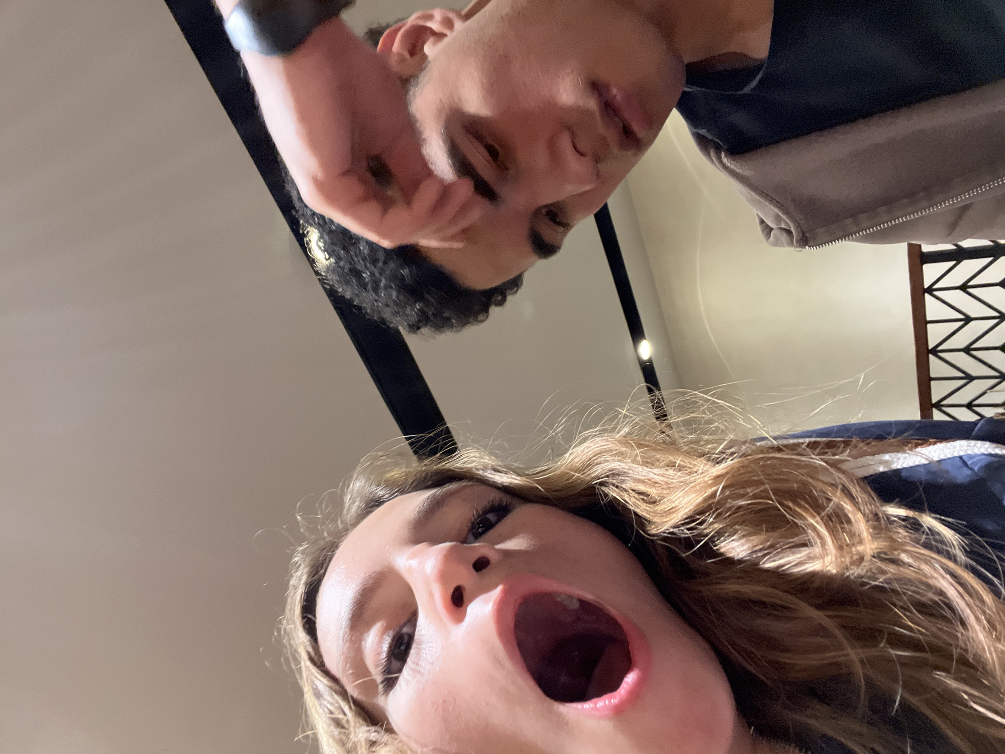 Two individuals are present in the image, which is taken from a low angle, looking upwards. The woman in the foreground has light brown hair and her mouth is open wide. The man behind has dark hair and is looking down at the camera, with his hand near his face. A dark object is visible on his wrist. The background suggests an indoor setting. This photo was not taken in Puebla, Mexico. There is not any way to get that information from the image.