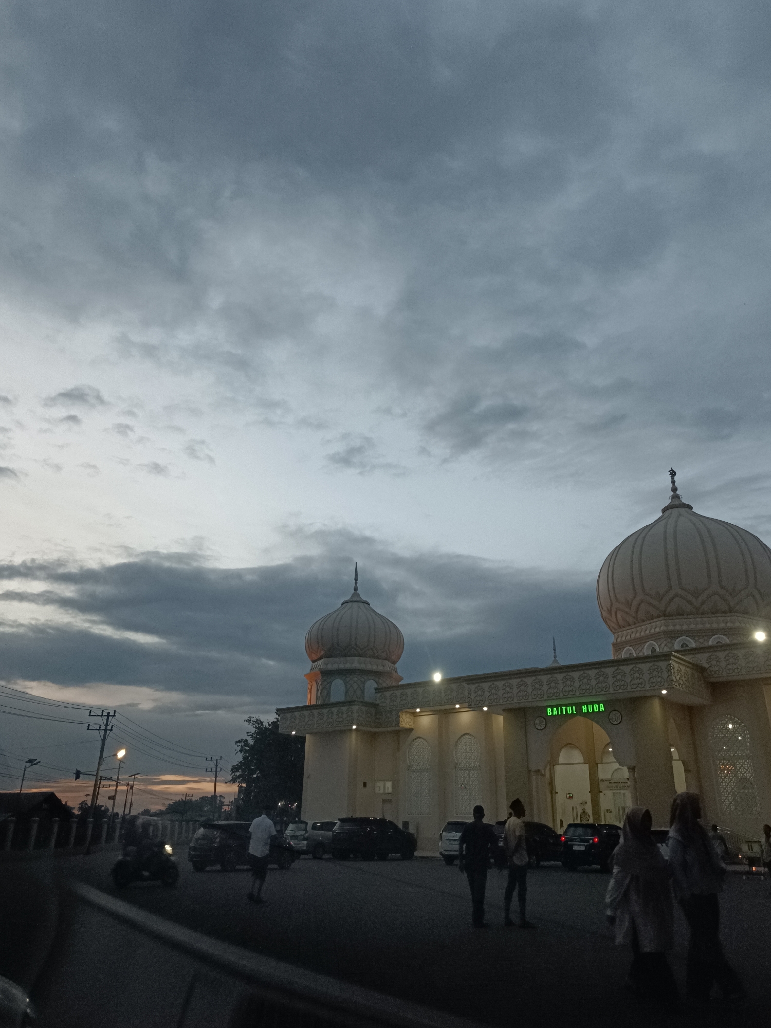 This media file captures a scene at dusk in front of a mosque named "BAITUL HUDA" in Mee Tanjong Usi, Indonesia. The sky is filled with dramatic, dark gray clouds, suggesting overcast or stormy weather. The mosque, an imposing structure with prominent domes and intricate architectural details, is illuminated by artificial lights, creating a warm glow against the dimming sky.
Several individuals are visible in the foreground and near the mosque entrance. Some are walking, while others appear to be standing and observing. A few cars are parked in front of the mosque, and a motorcycle is also present on the road. Power lines and streetlights are visible in the background, indicating an urban or semi-urban setting.
The overall mood of the image is serene yet somewhat dramatic due to the cloudy sky. The time of day is clearly indicated by the transition from daylight to evening, with the lights of the mosque and streetlights beginning to dominate the illumination. The presence of people suggests that the mosque is a place of gathering or prayer, and the activity appears to be the end of the day's activities. The text "BAITUL HUDA" is prominently displayed in green neon letters above the mosque's entrance.