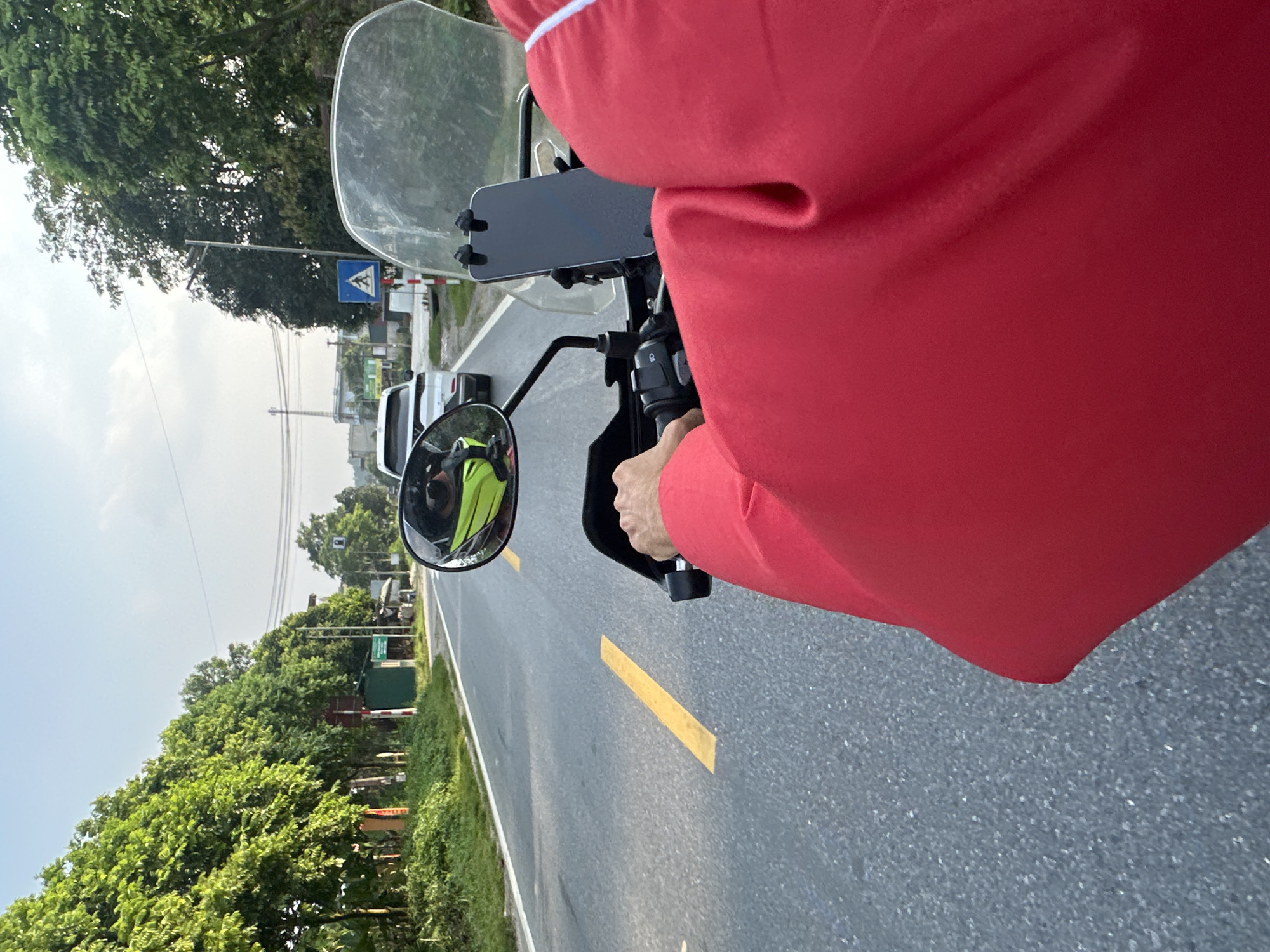 The image provides a first-person perspective from a person riding a motorbike along a paved road in Vị Xuyên Commune, Vietnam, during clear daylight hours. The rider, wearing a red long-sleeved top, has their right hand gripping the motorbike's throttle. A smartphone is mounted on the handlebars, situated just beneath a clear windshield. The rider's reflection, clad in a bright yellow or green garment, is visible in the left rearview mirror.
The asphalt road features a prominent solid yellow line on the left side and a white dashed line on the right, guiding traffic flow. Both sides of the road are bordered by abundant green trees and lush foliage, indicating a verdant, possibly rural or semi-urban setting. In the distance, several cars can be seen on the road.
Notable roadside features include a blue square sign displaying a white pedestrian crossing symbol on the left side of the road. Further down, a red and white striped barrier, characteristic of a railway crossing, is visible, indicating an upcoming intersection with a train track. Utility poles and overhead wires are also part of the landscape. The bright, clear sky suggests sunny weather, offering good visibility for travel. The scene captures a moment of peaceful transit through the Vietnamese countryside.