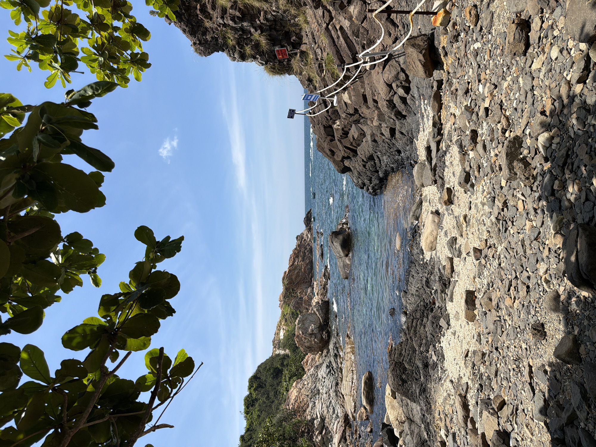 The image depicts a coastal scene with a rocky beach and clear blue water. Lush green foliage frames the top of the image, suggesting a tropical or subtropical environment. The beach is covered with various sizes of stones and pebbles, with a section of lighter colored sand in the middle ground. Dark, rugged rock formations line the right side of the scene, with a white rope railing guiding a path along the edge. Several blue and white signs are visible attached to poles along the railing, and a red sign is affixed to the rock face further up. The water is calm, with small waves lapping at the shore. In the distance, more rocky hills covered in vegetation rise from the sea, and a few small boats can be seen on the water. The sky is a bright blue with a few scattered white clouds. The overall impression is of a natural, somewhat rugged coastline. The location is identified as Phú Lương, Vietnam.
