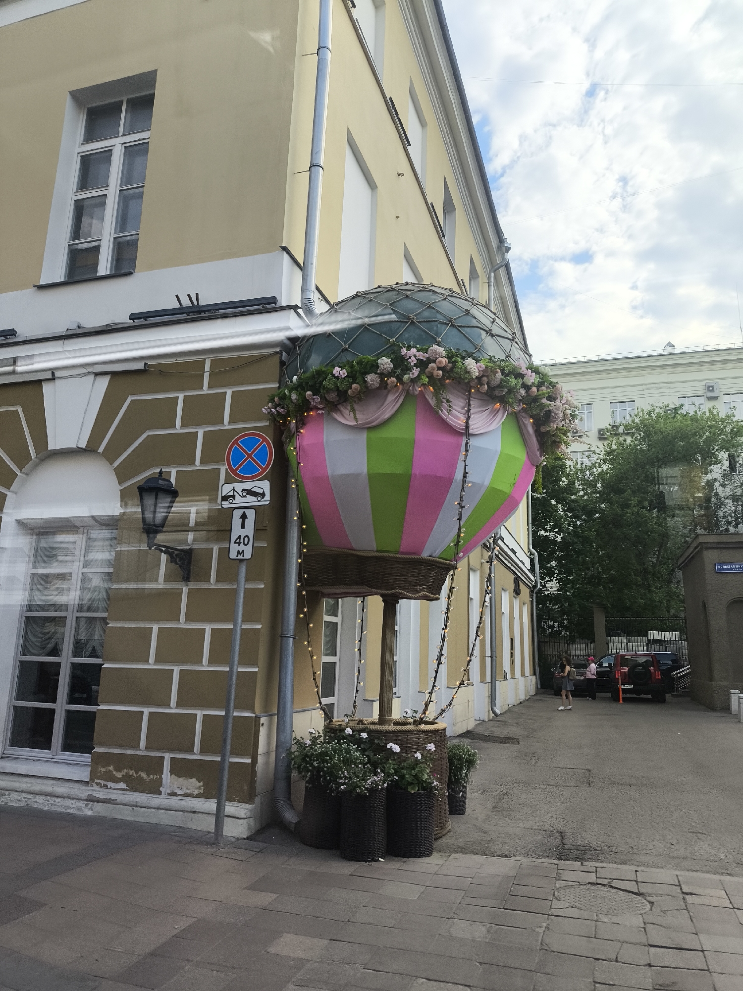 The image shows a colorful hot air balloon decoration attached to the corner of a light-yellow building in an urban setting. The balloon is decorated with flowers and has a basket underneath, resembling a real hot air balloon. There is a street sign indicating "No Stopping" and a speed limit of 40, with "M" underneath, suggesting the limit is in meters. The building has windows and a brick-like facade with white trim. In the background, there is a street with parked cars and pedestrians. The location is Unknown, Unknown.