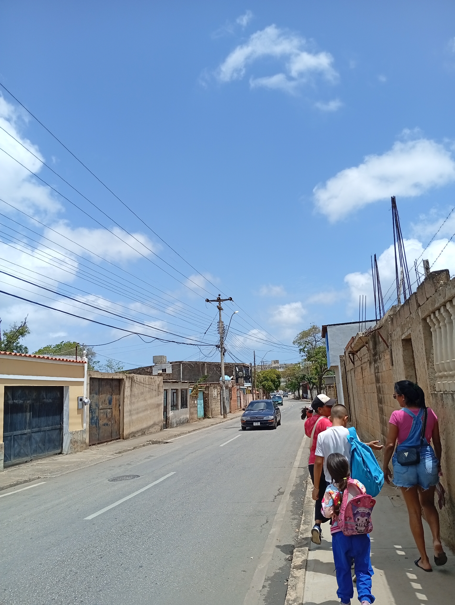 A group of people, including two adults and three children, walk along a sidewalk next to a street in Juan Griego, Venezuela. The group is walking away from the camera and towards the background of the image. The adults are a woman wearing denim overalls and a pink shirt, and another adult wearing a white shirt and a black and white hat. The children are two boys and a girl, all wearing backpacks. The street is paved and appears to be in good condition, with some cars driving in the distance. The buildings lining the street are mostly one and two stories high and appear to be made of concrete or stucco. There are power lines running overhead, typical of urban areas. The sky is blue with some scattered white clouds. The overall scene depicts a sunny day in a residential or commercial area of Juan Griego.
