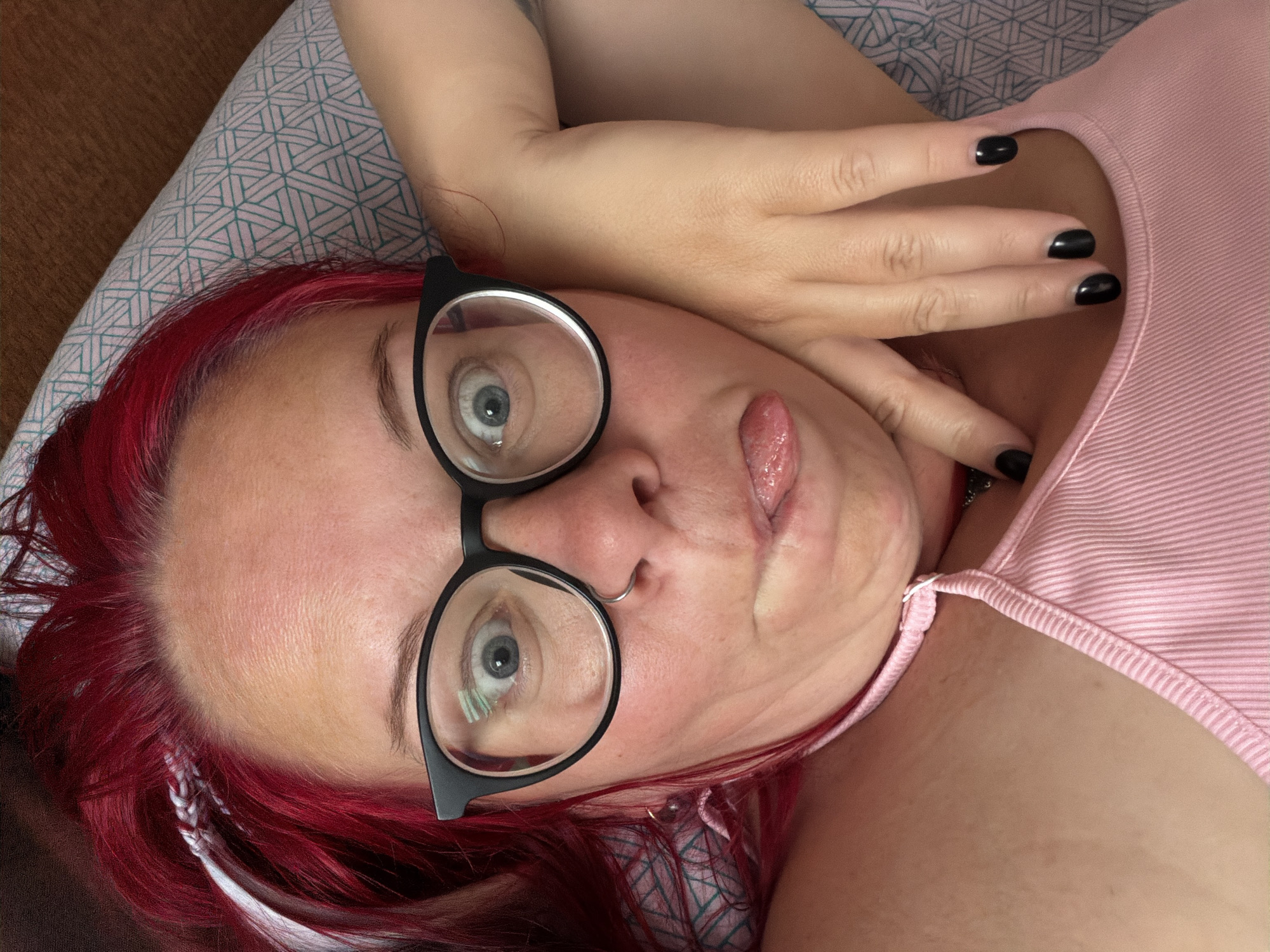 In a close-up, overhead shot, a woman with vibrant red hair and black-rimmed glasses makes a playful face, sticking out her tongue. She wears a pink tank top and has black nail polish. Her left hand rests on her cheek, and her right arm is bent, with her hand near her neck. The background features a pillow with a geometric pattern in shades of light blue and white, and a brown surface. This scene does not depict any recognizable location in Chaikovsky, Russia.