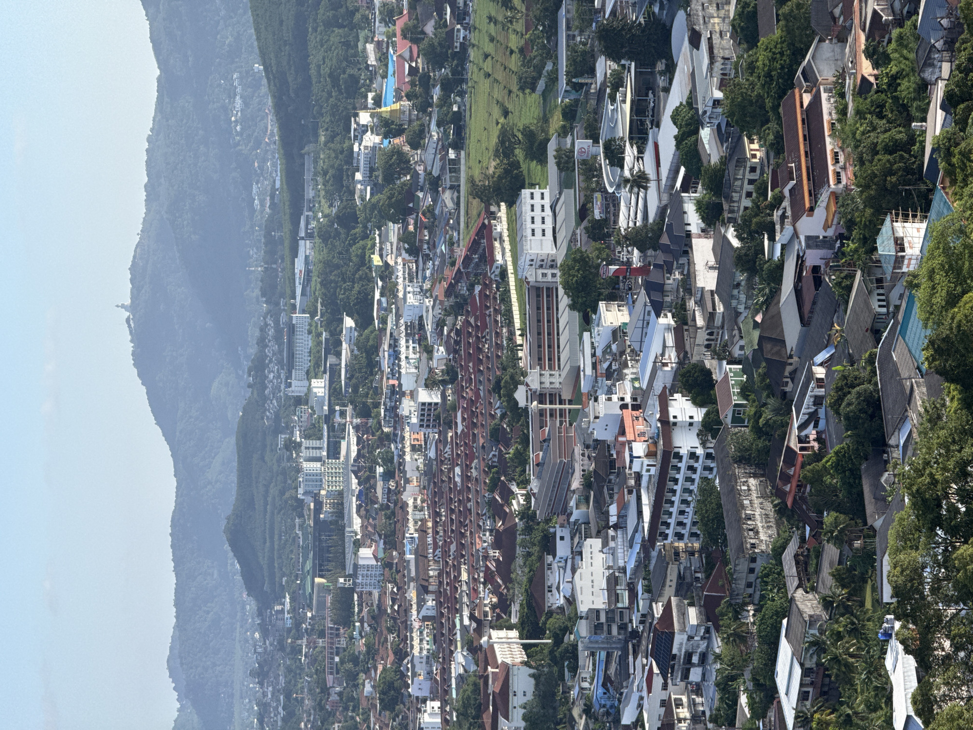 This is an aerial photograph of a densely populated urban area, likely within the vicinity of Ratsada, Thailand, characterized by a mix of residential buildings, commercial structures, and natural vegetation. The scene is dominated by rows of houses with red-tiled roofs, indicative of a suburban or densely packed residential neighborhood. Among these, larger, multi-story buildings, possibly apartment complexes or commercial establishments, are interspersed throughout the landscape.
In the background, lush green hills rise, suggesting a mountainous or hilly terrain surrounding the city. The sky is clear and pale blue, with no visible clouds. The lighting indicates it is daytime, likely mid-morning or late afternoon, given the soft shadows. There are no people or animals clearly visible in the image, and no discernible actions or interactions are taking place.
Several notable structures are visible. A prominent golden stupa, characteristic of Buddhist temples, stands out among the buildings on the right side of the frame. Further in the distance, on a hilltop, a large white statue, possibly of the Buddha, is visible, a common landmark in Thailand. In the mid-ground, a large building with a distinctive green roof structure might be a stadium or a large civic building. Commercial signage, such as a "TOYOTA" sign, is visible on a building, indicating commercial activity. The overall impression is one of a sprawling, established urban area integrated with its natural surroundings.