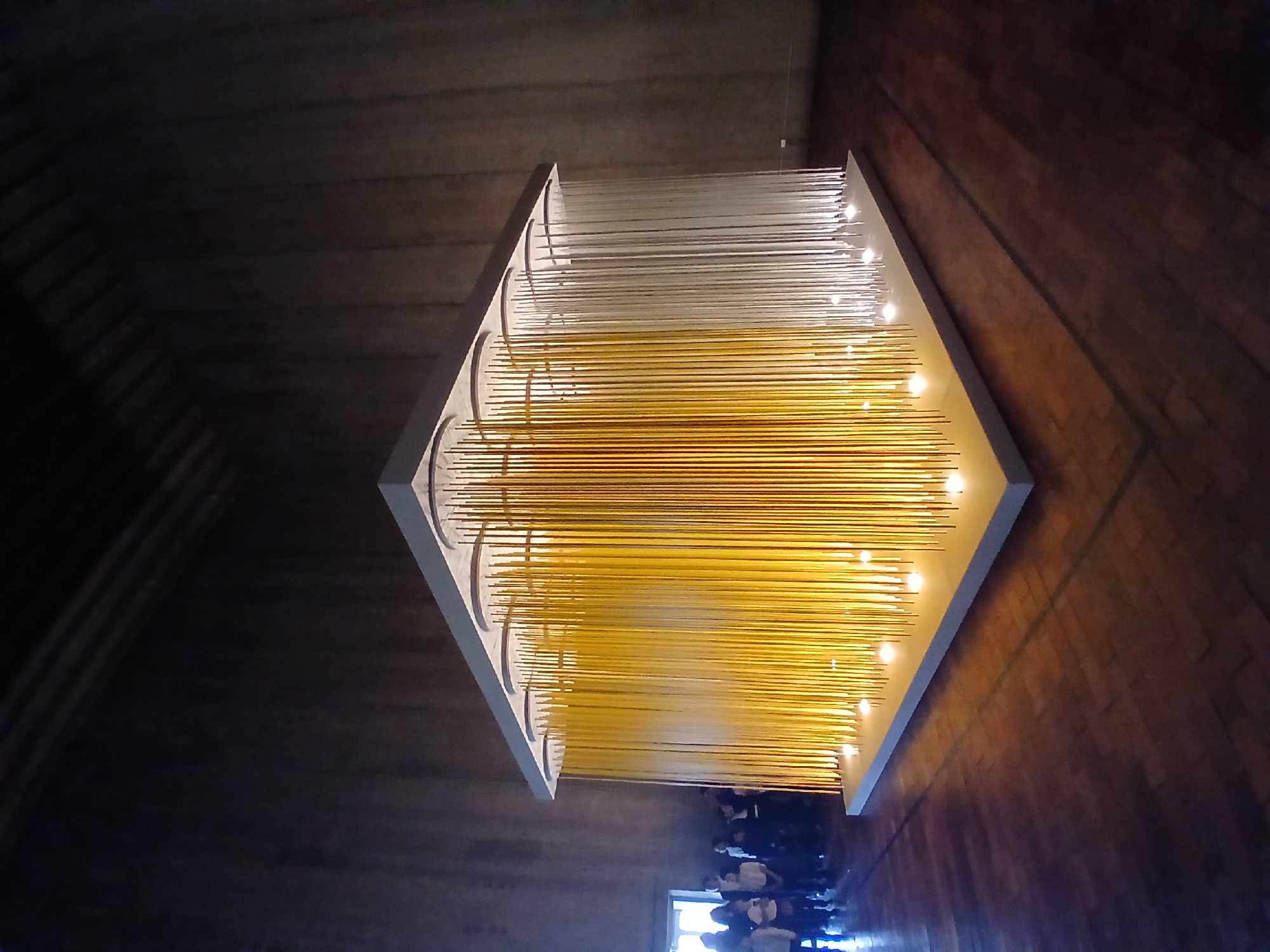 The image shows a large, square chandelier hanging from the ceiling of a building in Ciudad Bolivar, Venezuela. The chandelier consists of a white frame with rows of vertical, cylindrical rods. The top portion of the rods is white, transitioning to a yellow color in the lower portion. Small lights are spaced along the bottom edge of the frame. The ceiling is dark, and the walls of the building appear to be constructed of dark wood or stone. A group of people is visible in the bottom center of the frame. They appear to be looking up at the chandelier. A window is visible in the bottom left corner.