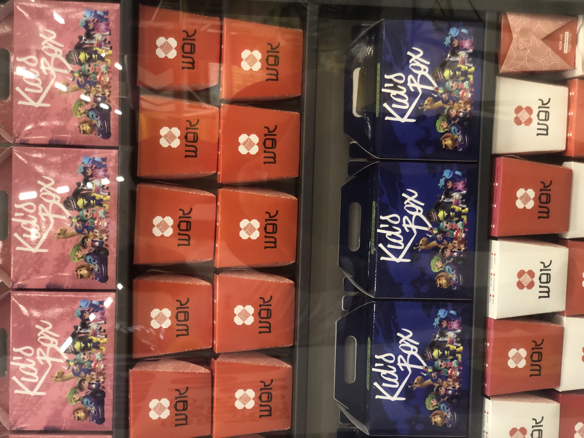 The image displays a retail environment, likely a supermarket or a dedicated food establishment, showcasing various food packaging. The primary subjects are rows of brightly colored boxes and containers.
In the top row, three pink boxes, labeled "Kid's Box" in white cursive lettering, are visible. These boxes feature illustrations of animated characters, including cartoon animals and anthropomorphic figures, with some characters riding in what appears to be a small vehicle.
Below the pink boxes, a shelf contains an array of red and white containers. The red containers are predominantly visible, each featuring a white logo that resembles a stylized flower or knot, with the word "WOK" printed below it in black lettering. Some of these red containers have a patterned texture on the sides.
The bottom row features darker blue boxes, also labeled "Kid's Box" in white lettering. These boxes display a similar illustration style to the pink boxes, with a group of animated characters.
One red container on the far right of the bottom shelf, partially visible, has the text "wok.uz" and a phone number printed on it. This detail suggests a connection to a specific brand or establishment operating in Uzbekistan, likely Tashkent, given the ".uz" domain. The overall impression is a display of children's meal packaging and takeaway food containers. The lighting suggests an indoor setting with artificial illumination.
