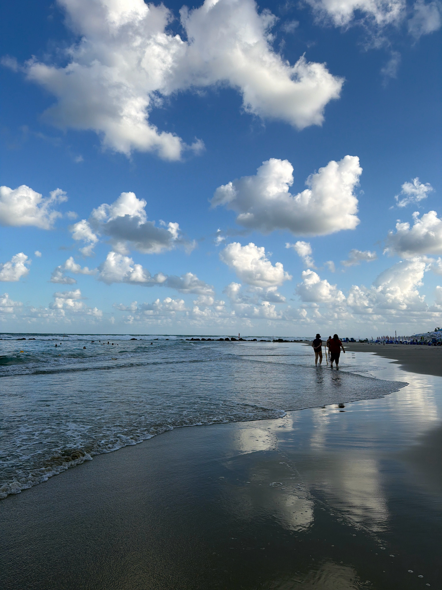 The image captures a serene beach scene in Bat Yam, Israel, under a bright blue sky dotted with fluffy white clouds. The foreground is dominated by the wet sand of the beach, reflecting the sky and clouds like a mirror. A gentle wave is seen lapping at the shore, creating a foamy edge. In the middle ground, two figures, silhouetted against the light, are walking along the water's edge. The sea stretches out to the horizon, with a few distant swimmers visible. Further back, along the shoreline, a row of beach chairs and umbrellas suggests a populated area. The overall atmosphere is peaceful and idyllic, a typical day at the beach.