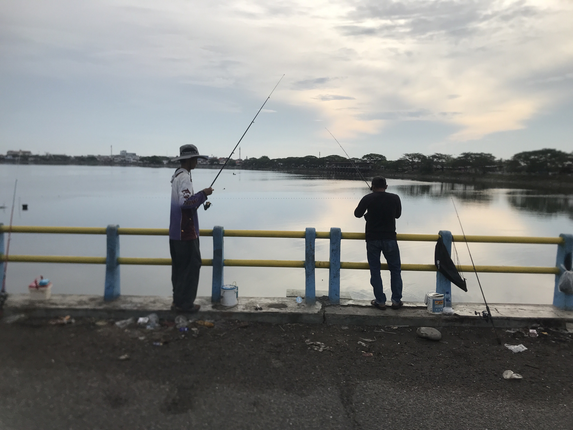 The image depicts two individuals fishing from a concrete pier at a body of water, likely a lake or bay, in Lhokseumawe, Indonesia. The sky is overcast with subtle hues of pink and orange near the horizon, suggesting either sunrise or sunset. A distant cityscape with buildings and trees lines the far bank of the water.
On the pier, the person on the left wears a wide-brimmed hat, a long-sleeved shirt with purple and white accents, and dark pants. They are holding a fishing rod. To their right, another person is dressed in a dark long-sleeved shirt and blue jeans, also holding a fishing rod. A yellow railing with blue posts runs along the edge of the pier, separating it from the water. Various items are scattered on the pier surface, including buckets, a fishing reel, and some debris. To the left, a small boat with a red and white top is visible on the water. The foreground shows a paved surface, likely a road, with more debris. The overall scene conveys a calm and somewhat mundane atmosphere.