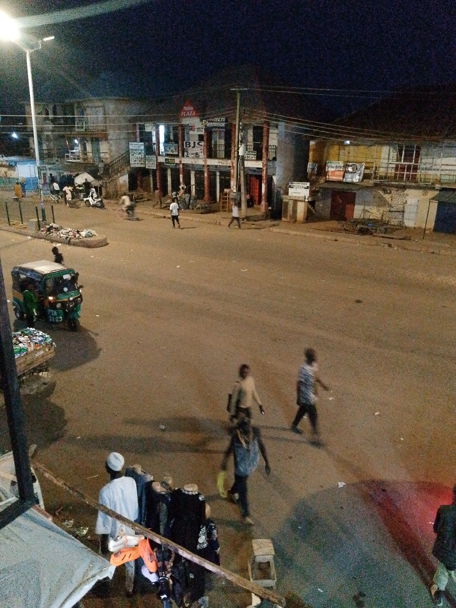The image depicts a nighttime street scene in Bukuru, Nigeria. A street light illuminates a section of the asphalt road, revealing several people and vehicles. In the foreground, a man in a white robe and cap stands facing away from the camera, near a display of clothing and footwear. To his left, a three-wheeled auto-rickshaw, green and white in color with the license plate "PTA 9123", is parked on the road.
Further down the road, several individuals are walking. One man in a light-colored shirt is walking towards the right, while another in a blue and white striped shirt is also walking in the same direction. Another person in a denim shirt and dark pants is walking towards the left. In the background, a two-story building with balconies and shops is visible, with signs including "Mahia PLAZA" and "BJC". Several other people are present in front of the shops, some appearing to be interacting or standing around. There are also motorcycles and other vehicles parked or moving on the street. Trash is piled on the side of the road near the street light. The overall environment suggests a commercial area that is active at night.
