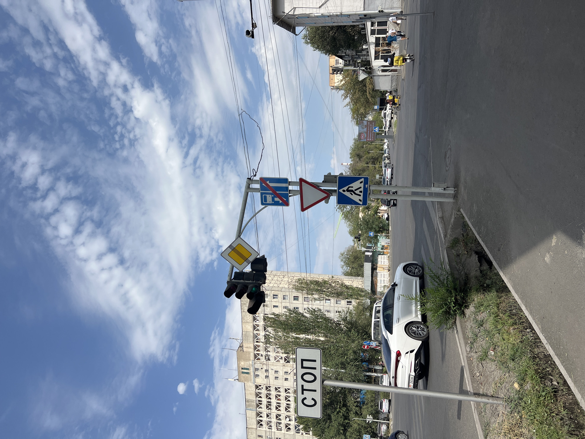 The image shows a street scene in Bishkek, Kyrgyzstan, likely at an intersection. In the foreground, a white car is driving across the intersection. A "STOP" sign is visible on the left side of the road, along with traffic lights. Further in the background, there is a large, multi-story building with a distinctive architectural pattern. The sky is blue with scattered clouds. The overall scene depicts an urban environment with typical street infrastructure.