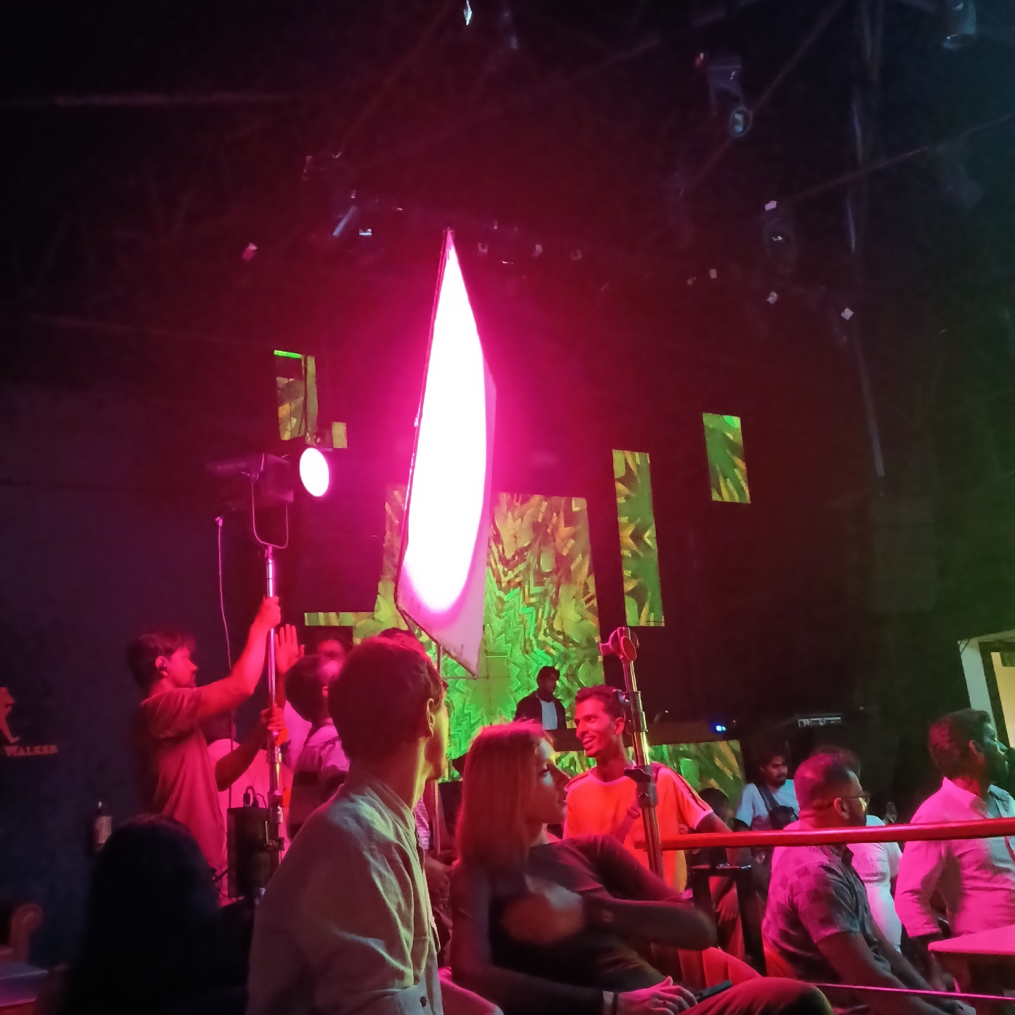 Captured in the city of Sinquerim, India, the scene unfolds under a vibrant red and green lighting scheme. A group of people are observed, some seated and some standing. Among them, two individuals are distinctly highlighted by a bright light panel, possibly for filming or photography purposes. The backdrop consists of vertically oriented rectangular screens displaying a green, abstract pattern. Overhead, a grid-like structure hangs above the scene, supporting additional lighting fixtures. The location suggests a nightclub or event space.