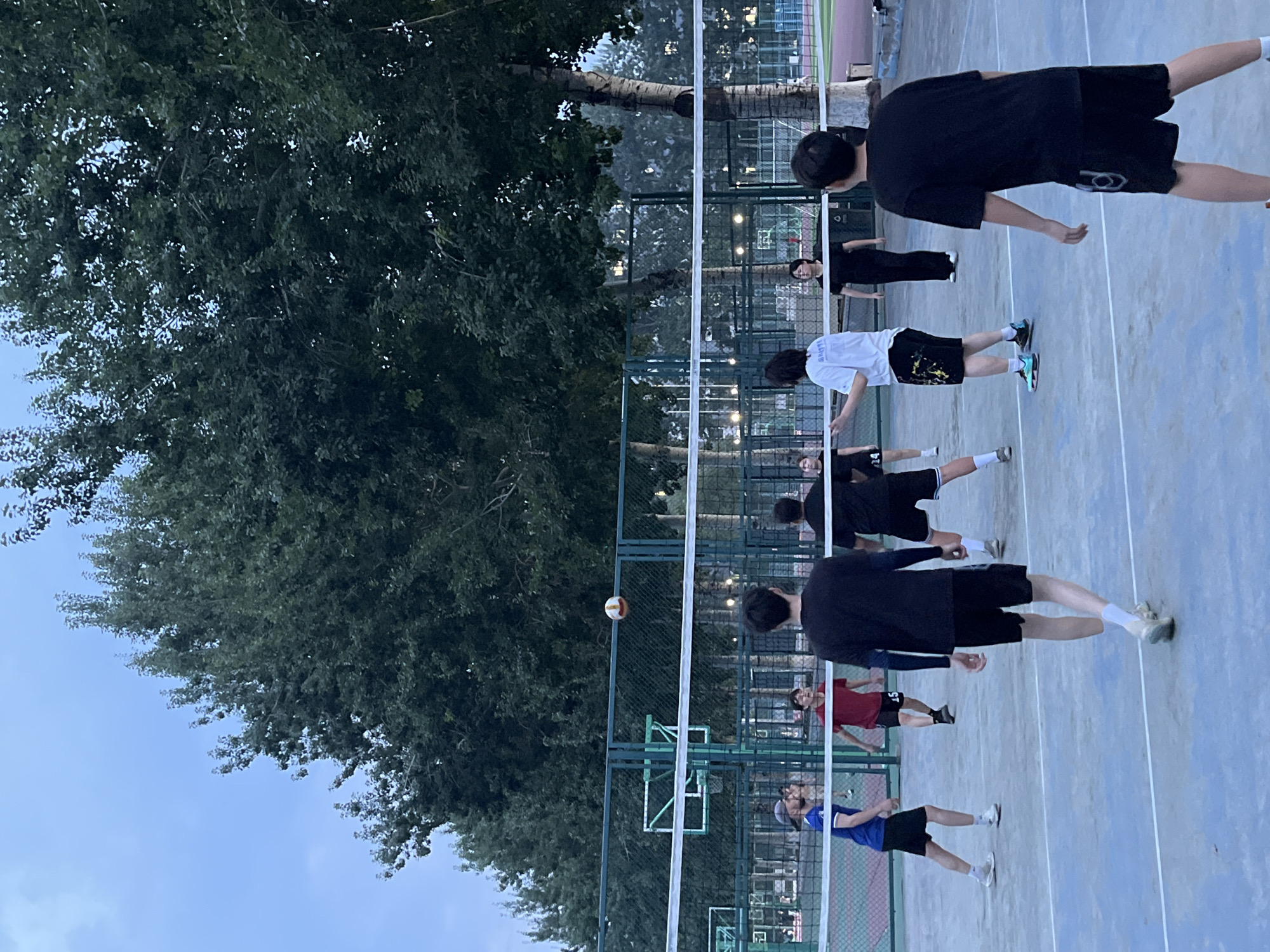 In the city of Daxing District, China, a group of people are playing volleyball on an outdoor court. The sky is overcast and the trees are dense with foliage. The volleyball court has a blue surface with white lines. A white net stretches across the court. On one side of the net, a person in a blue shirt and black shorts is in motion, with their body turned towards the net and their arms extended. To their right, a person in a red shirt and black shorts is visible. Further into the court, several other individuals are engaged in the game, with some looking towards the volleyball, which is suspended in the air above the net. The overall atmosphere appears casual and active, with people enjoying recreational activity in an outdoor setting.