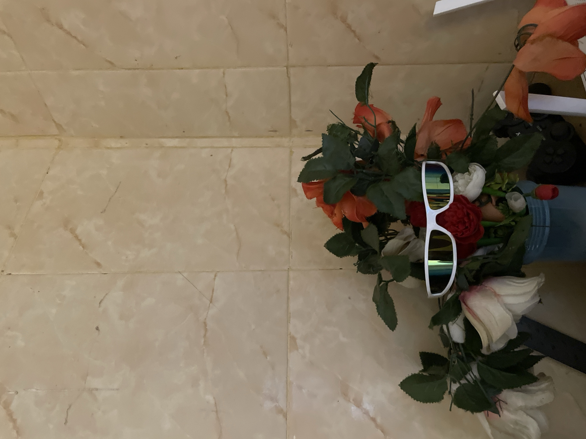 In Ado, Nigeria, a still life arrangement features a bouquet of artificial flowers, primarily in shades of orange, red, and white, placed in a blue vase. Positioned among the flowers are a pair of white, futuristic-style sunglasses with reflective lenses. The background consists of a tiled wall with a marble-like pattern.