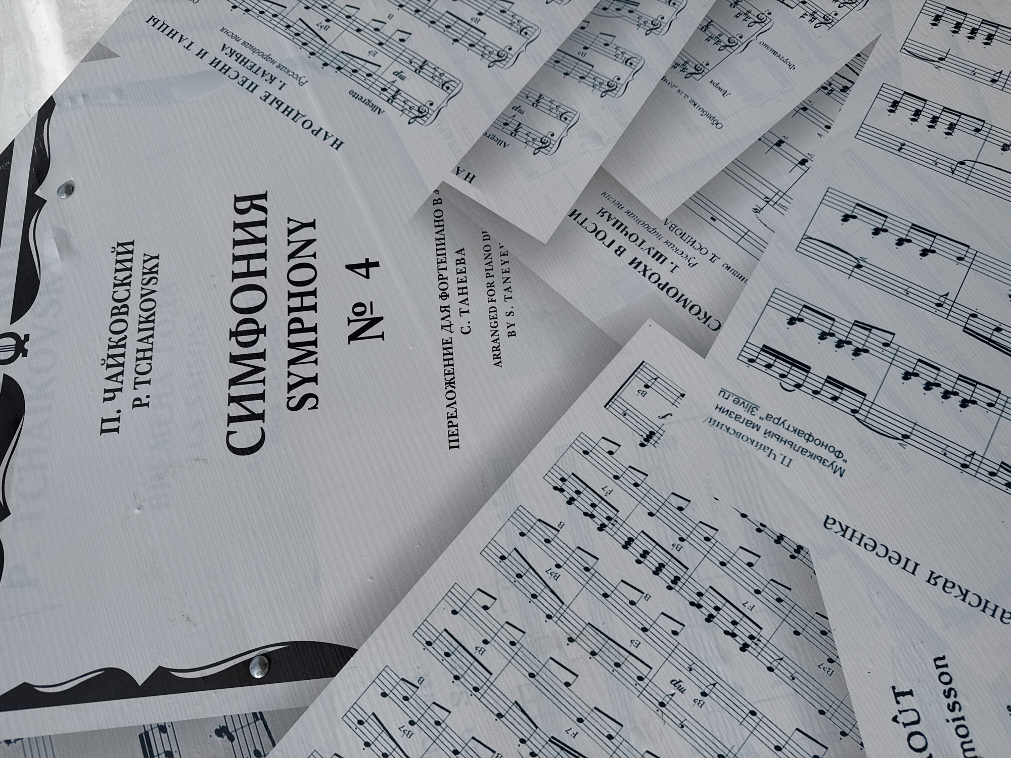 The image shows a collection of sheet music, seemingly scattered or layered on a surface. The music is in black and white and includes both musical notation and Cyrillic text. The title of one piece prominently displayed reads "СИМФОНИЯ SYMPHONY No. 4" by P. TCHAIKOVSKY. There are other songs called "СКОМОРОХИ В ГОСТИ" and "НАРОДНЫЕ ПЕСНИ И ТАНЦЫ". Other details include "ПЕРЕЛОЖЕНИЕ ДЛЯ ФОРТЕПИАНО В А" or "ARRANGED FOR PIANO DI". The presence of music notation with notes, clefs, and dynamic markings suggests the pages are intended for performance or study.