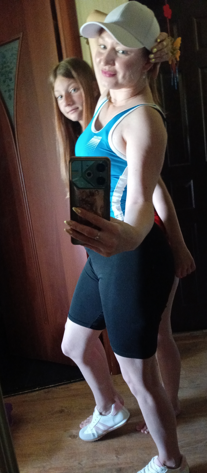 A woman with a white cap, blue and white tank top, and black shorts is taking a selfie in front of a mirror. Another woman is standing behind her. The woman taking the selfie is wearing white sneakers and has long, painted nails. The environment appears to be indoors, near a door. Location: Unknown, Unknown.