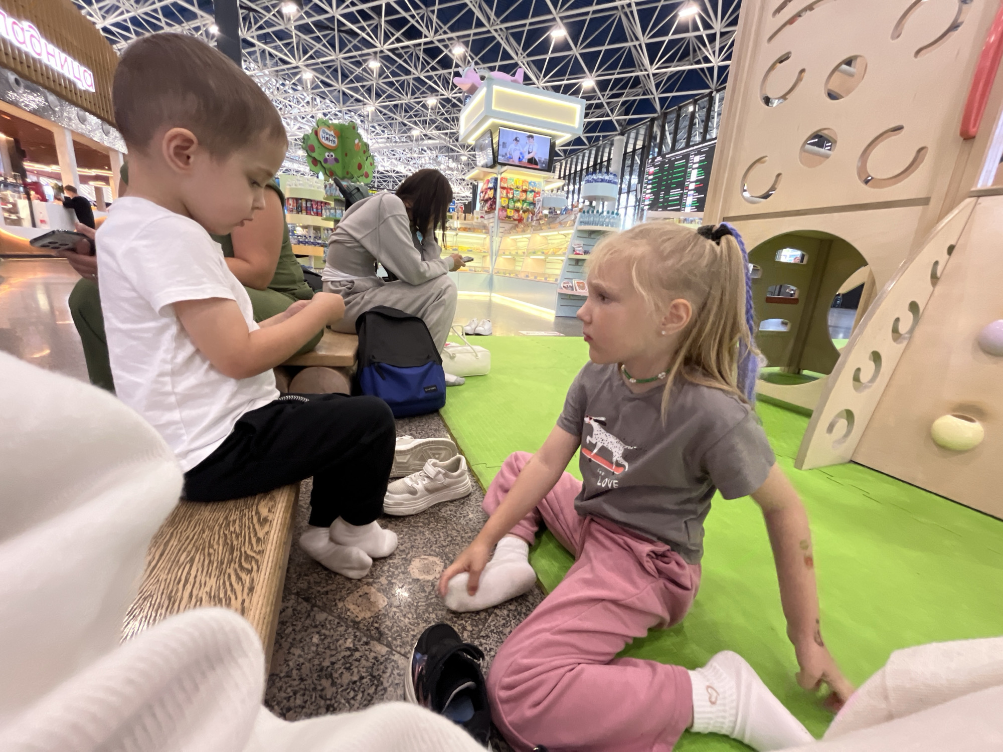 The image depicts an indoor play area, likely in an airport or shopping mall, with a focus on two young children. A boy with a white t-shirt and black pants sits on a wooden bench, looking down at his hands. Beside him, a girl in pink pants and a grey t-shirt with a dog graphic is sitting on a bright green mat, facing the boy and appearing to engage in conversation or play. Other individuals and parts of the play structure are visible in the background, suggesting a public, family-friendly environment. The architectural style of the ceiling suggests a modern, large-span building. Given the prompt, the location is specified as Sochi, Russia, implying this scene is within a facility in that city.