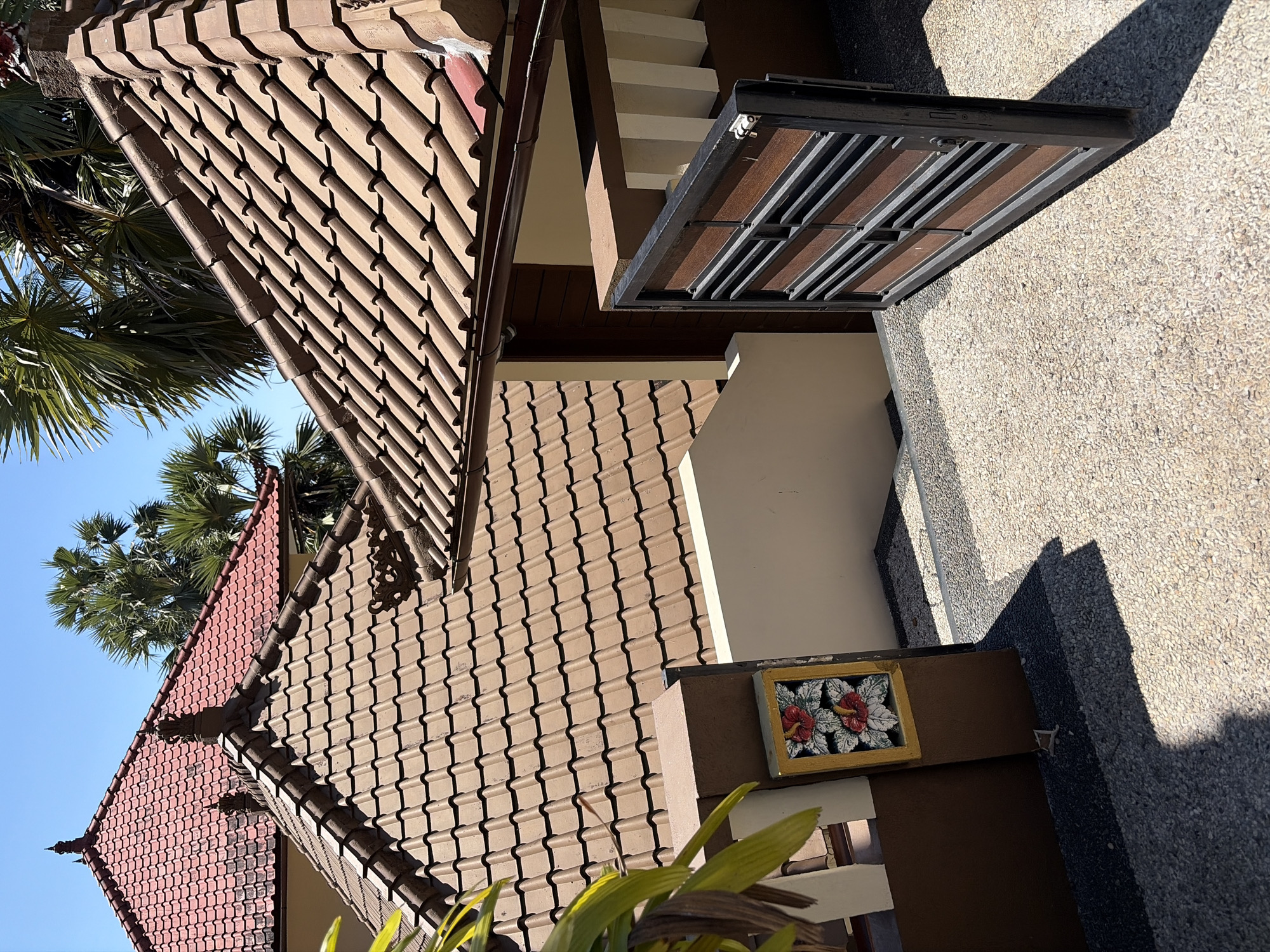 This is a medium shot, eye-level, outdoor photograph taken on a sunny day. The image depicts a courtyard entrance with an open gate. The gate is made of dark metal and has horizontal wooden slats. The courtyard is paved with small, light-colored pebbles. To the left of the gate, a brown pillar stands with a decorative tile featuring two red hibiscus flowers against a white and green background, framed by a yellow border. Beyond the pillar, a cream-colored wall rises, leading to a staircase.
The background features two buildings with terracotta tiled roofs, characteristic of Indonesian architecture. The roof in the foreground is brown, while the roof behind it is red. Lush green palm trees are visible above the roofs, suggesting a tropical location. The sky is a clear, bright blue. The image appears to have been taken in Abang, Indonesia, given the architectural style and vegetation. There are no people or animals visible in the frame, and no actions or interactions are taking place.