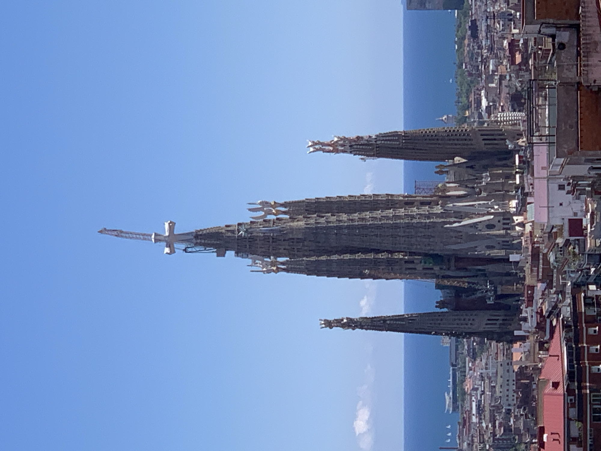 The image captures an upward view of the Sagrada Familia basilica in Barcelona, Spain, its intricate spires dominating the skyline. A large crane stands atop one of the spires, indicating ongoing construction. The basilica's detailed stonework is visible against a bright blue sky. The surrounding cityscape fades into the background, merging with the Mediterranean Sea on the horizon. Several smaller buildings with red roofs are visible closer to the frame.