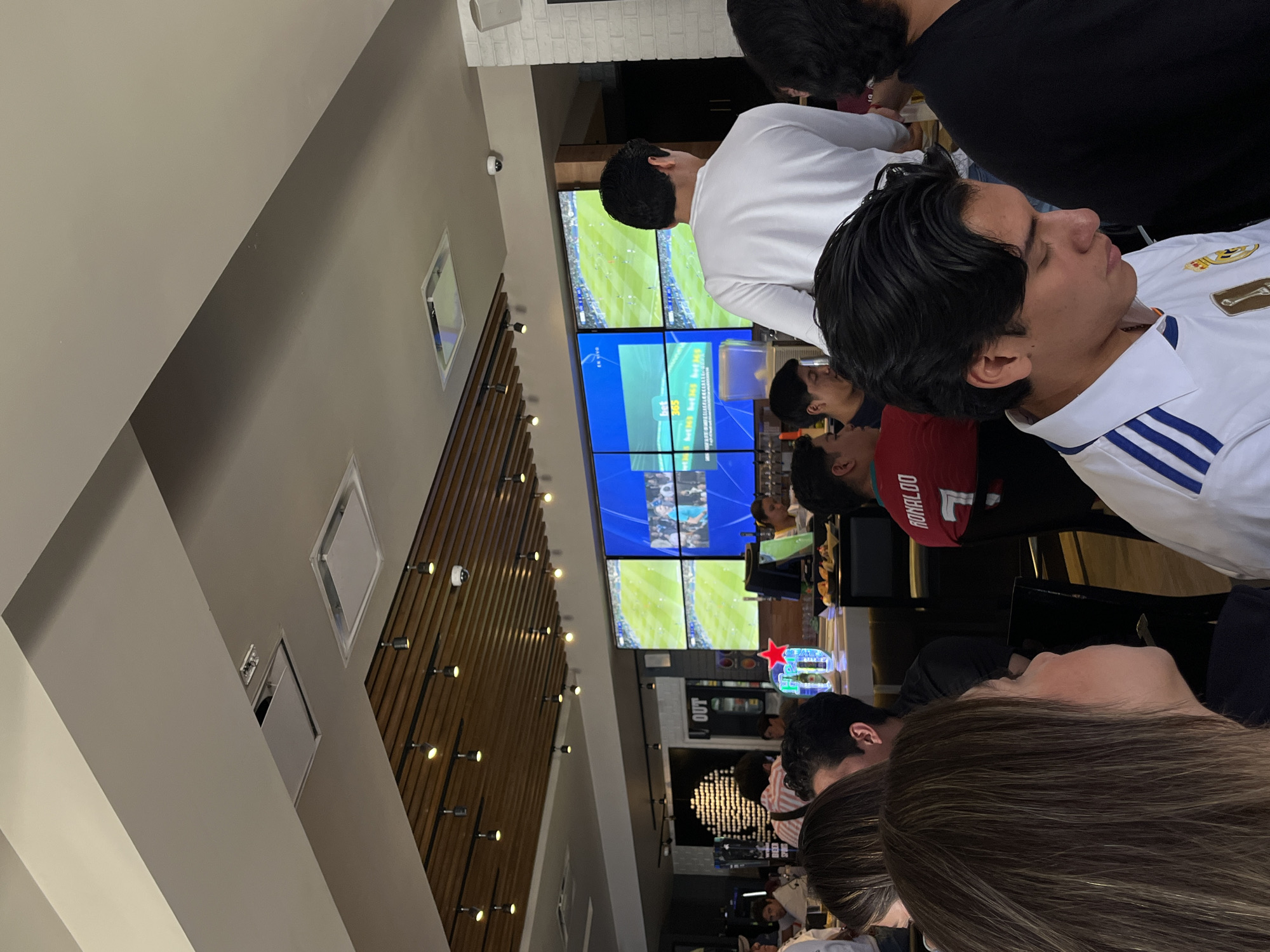 The image shows a group of people inside a bar or restaurant, likely in San Bernardino Tlaxcalancingo, Mexico, based on the prompt's location information. The group is watching a soccer game on a large screen composed of multiple monitors. People are wearing jerseys from different soccer teams. The interior of the establishment features wooden paneling and lighting.