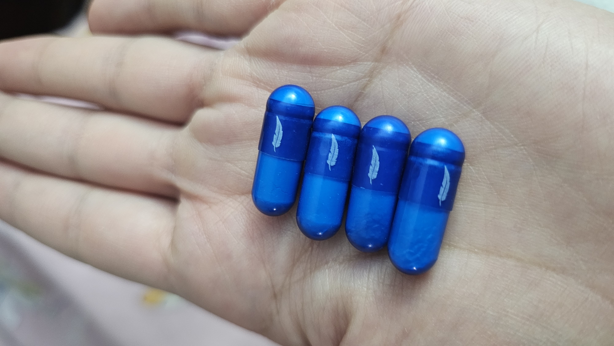 A close-up shot depicts four blue capsules, each adorned with a white feather design, resting in the palm of a hand. The capsules have a glossy finish and a two-toned color scheme, with a lighter blue shade on one end. The hand is light-skinned, with visible creases and lines. The background is slightly blurred, revealing a bed with patterned sheets. The location is unknown, but the city mentioned is San Bernardino Tlaxcalancingo, Mexico.