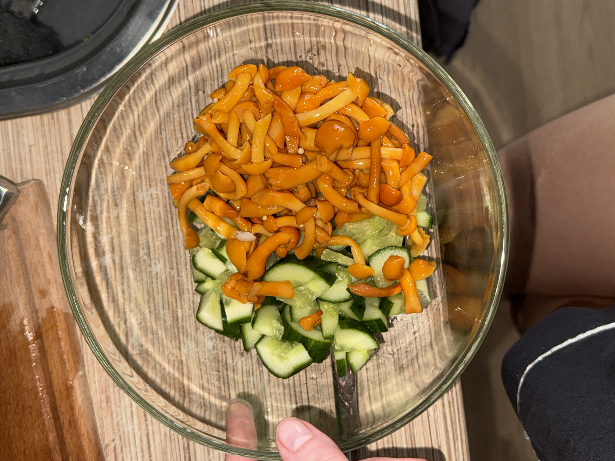 A clear glass bowl containing chopped cucumbers and bright orange enoki mushrooms sits on a wooden surface. The bowl is situated near a wooden cutting board and a dark metal object. A person's left hand is visible on the left side of the bowl, with fingernails that appear clean and manicured. The setting appears to be a kitchen or dining area. The location is not specified in Perm, Russia.