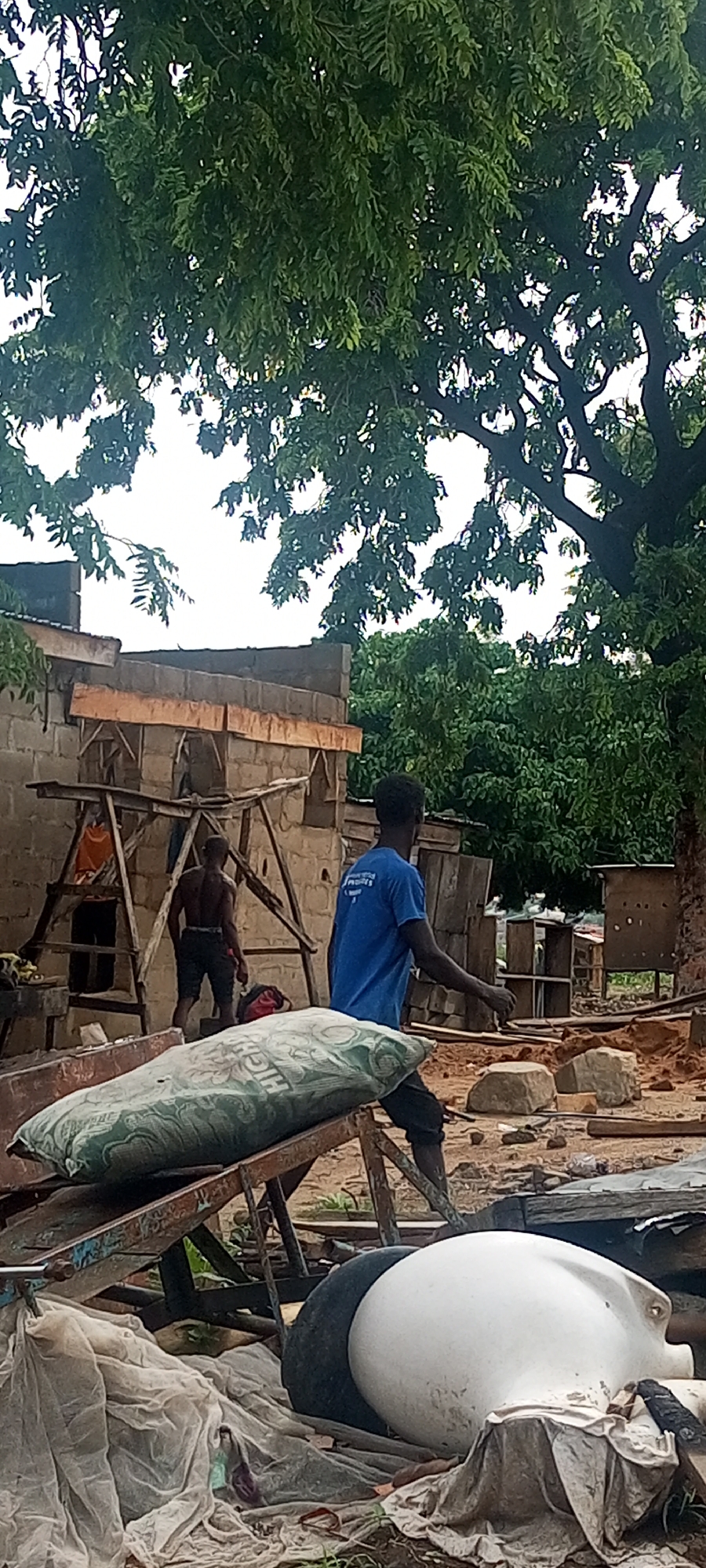 In Zango-ndaji, Nigeria, a construction site shows unfinished brick structures under the cover of leafy trees. Two shirtless men work on a makeshift scaffolding, while a third man wearing a blue shirt walks away from the structure. Discarded construction material, including bags of cement, tires, and debris, is in the foreground.