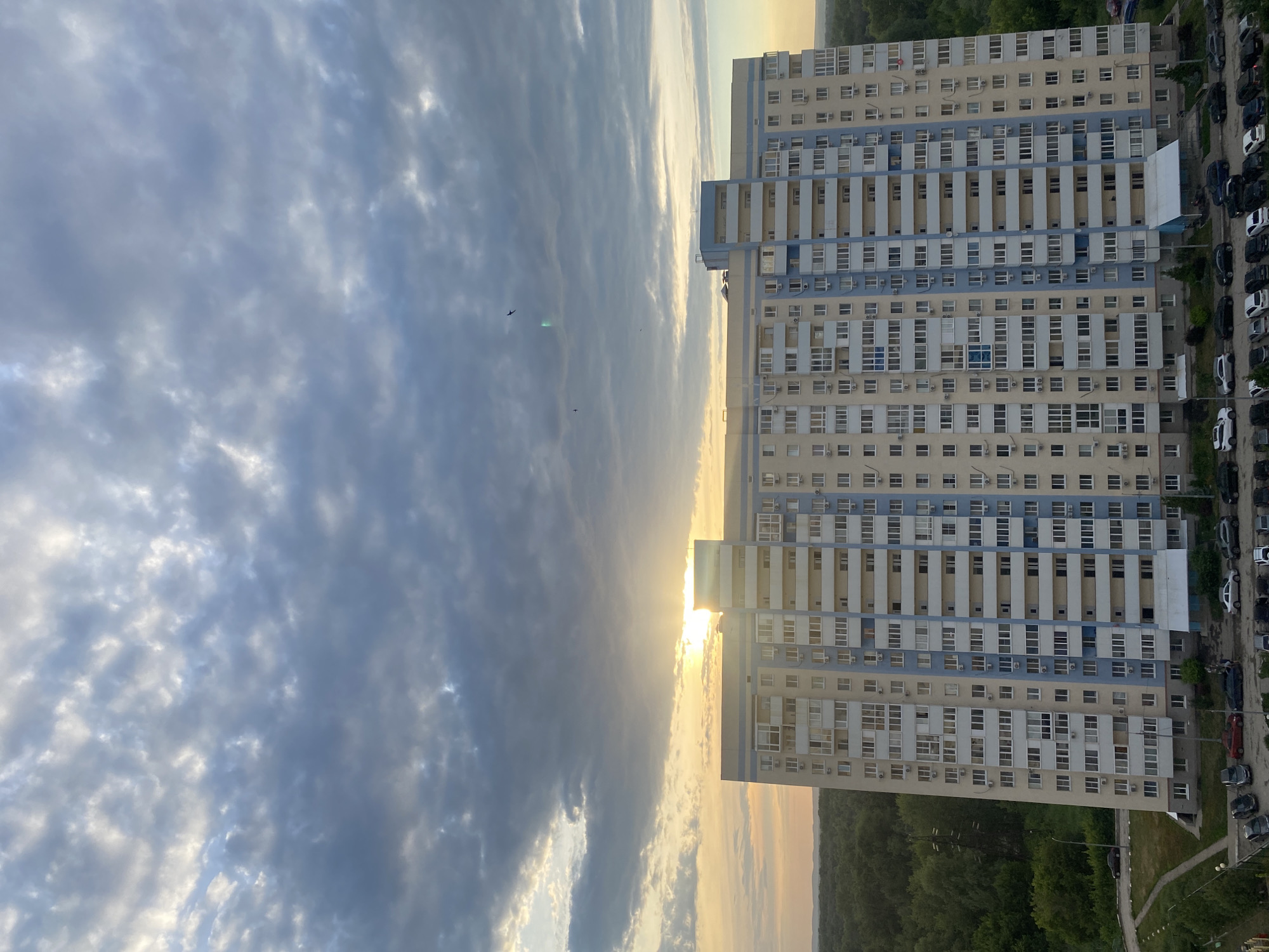 This is a vertical, eye-level, outdoor shot of a residential building in Novokuybyshevsk, Russia, during sunset. The building, which appears to be a high-rise apartment complex, dominates the right side of the frame. It is composed of numerous windows and balconies, with a pale yellow and light blue facade. The building is densely packed with many units, suggesting a large number of residents. To the right of the building, a line of parked cars is visible, indicating a residential area. The left side of the image shows a vast expanse of clouds, with the sun setting on the horizon, casting a warm, golden light. The sky is a mixture of dark and light clouds, with streaks of sunlight breaking through. A few birds can be seen in the sky. The overall mood is serene and peaceful, with the warm hues of the sunset creating a tranquil atmosphere. The location cues of Novokuybyshevsk, Russia, are provided externally and are not discernible from the image itself. There is no visible text in the image.