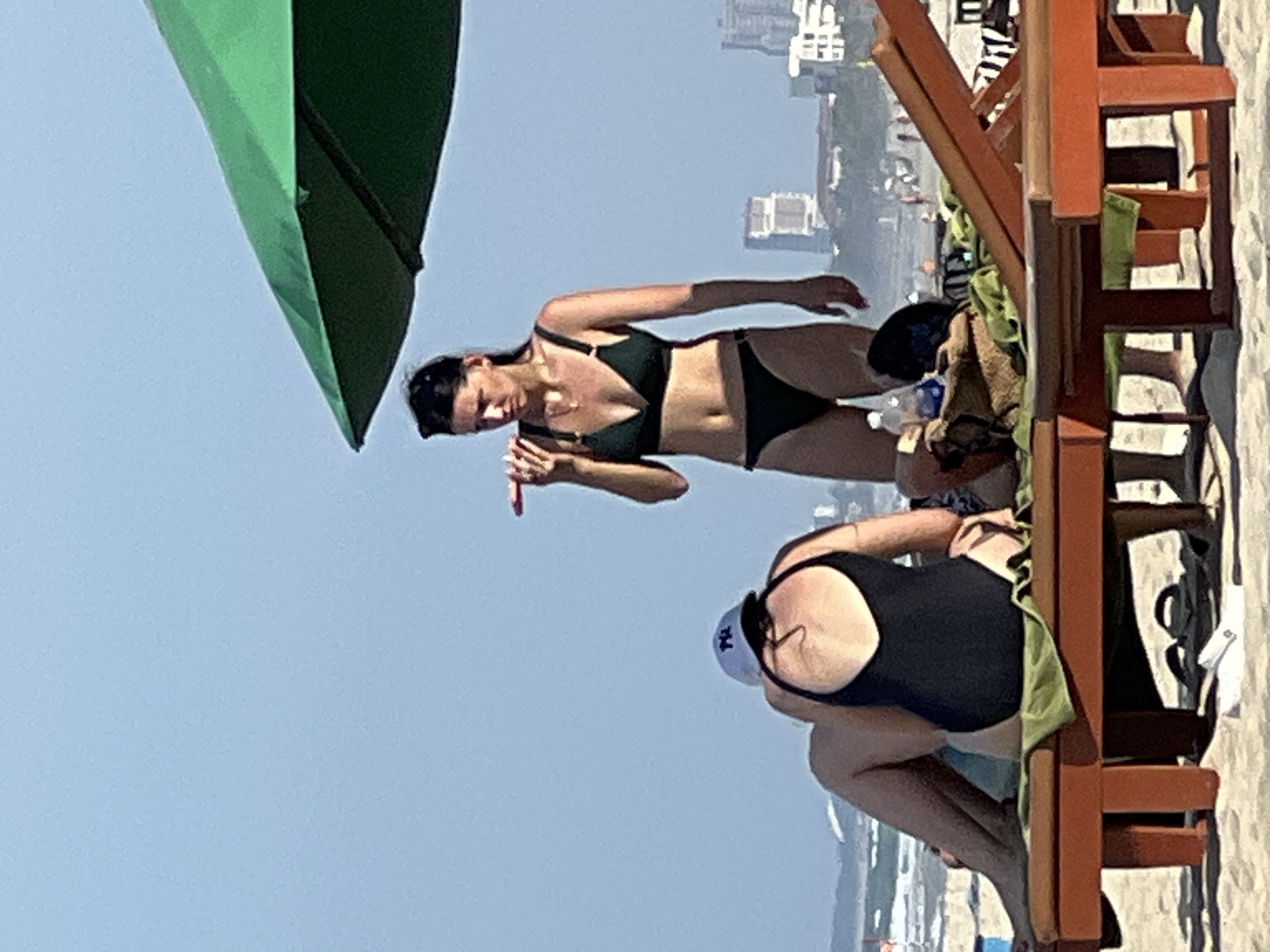 The image depicts a beach scene in Đà Nẵng, Vietnam. In the foreground, two individuals are seated on a beach chair. One person, wearing a black one-piece swimsuit and a white baseball cap, is facing away from the camera, showing their back. The other person, partially visible, is also seated. In the mid-ground, a woman in a black bikini stands and appears to be holding a cigarette or similar object to her mouth. She is looking to her left. Behind the individuals, a green beach umbrella provides shade. The background shows a hazy view of buildings and the ocean, indicative of an urban coastline. The sand and beach chairs suggest a leisure setting.