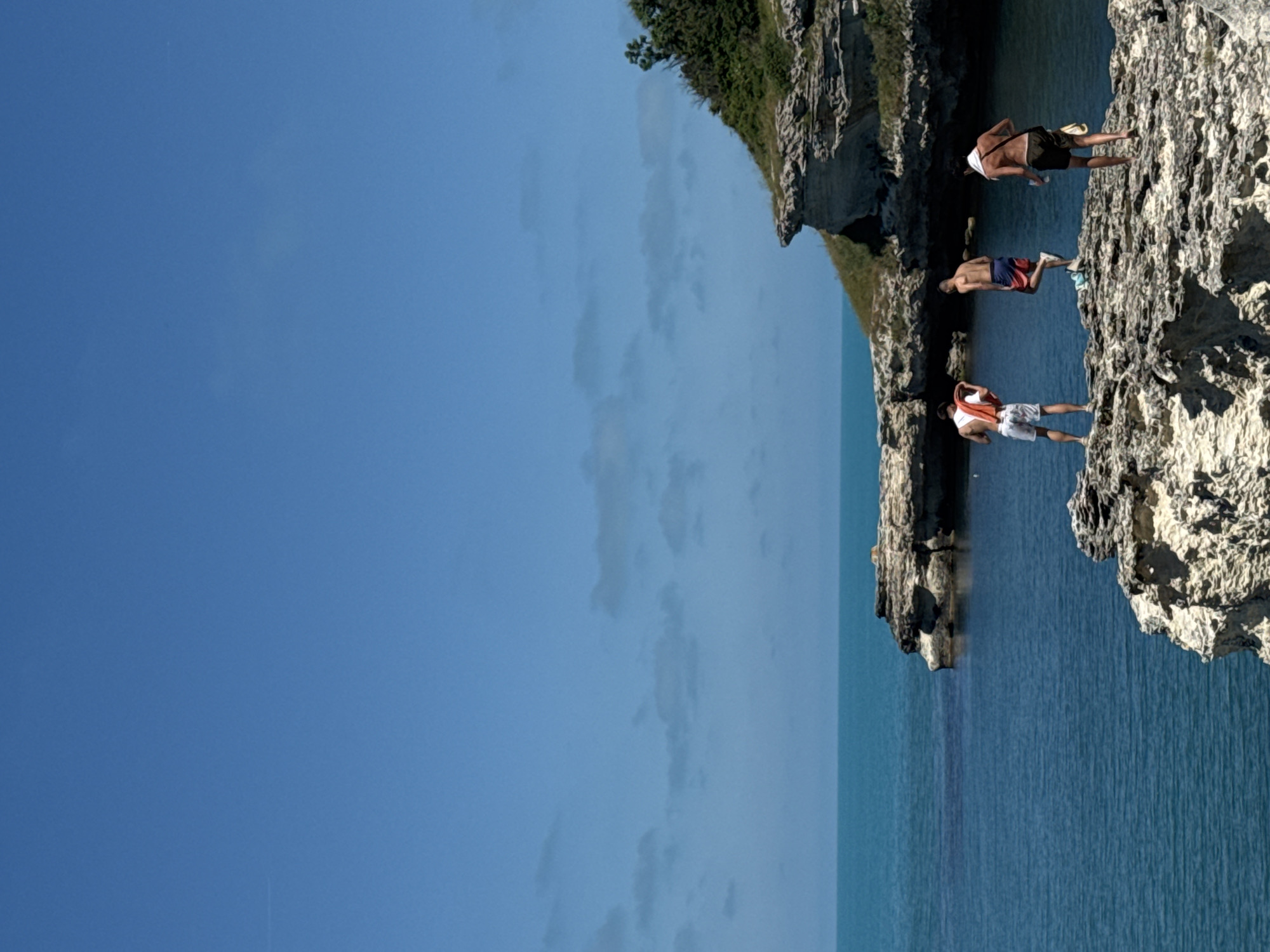 The image depicts three young men on a rocky cliff overlooking the Black Sea near Tsarevo, Bulgaria. The sky is a clear, bright blue with a few wispy clouds. The sea is a deep turquoise, calm and inviting. The men are dressed in swim trunks, and one is holding a towel. They appear to be enjoying a summer day at the beach, with the rugged coastline providing a dramatic backdrop. The rocks are rough and uneven, with patches of sparse green vegetation clinging to them. The overall impression is one of natural beauty and outdoor recreation.