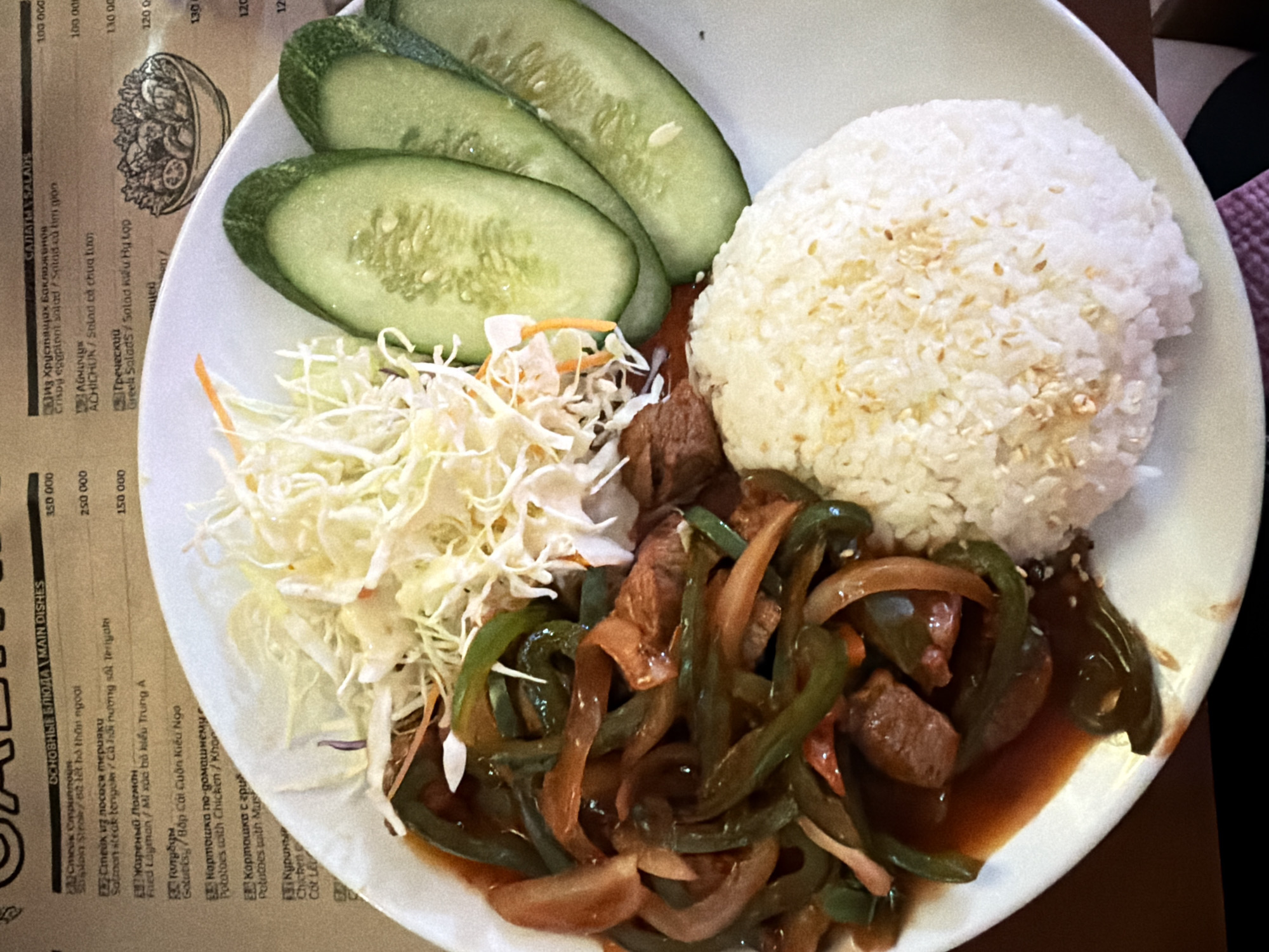 The image shows a plate of food served on a white plate. The plate contains a mound of white rice sprinkled with sesame seeds, a side of shredded cabbage and carrots, several slices of cucumber, and a portion of stir-fried meat and bell peppers in a dark sauce. The food appears to be a main dish, likely from an Asian cuisine.
In the background, a menu is visible with text in both Russian and English, indicating that the restaurant offers a variety of dishes, including salads, salmon steak, fried lagman, and chicken. Some prices are also visible on the menu. The menu also includes Vietnamese phrases, suggesting the location is in Vietnam, specifically Nha Trang Ward.
The setting appears to be an indoor restaurant or eatery. The lighting suggests it could be daytime or evening. There are no people visible in the image, and the focus is entirely on the food and the accompanying menu. The overall impression is of a well-presented and appetizing meal.