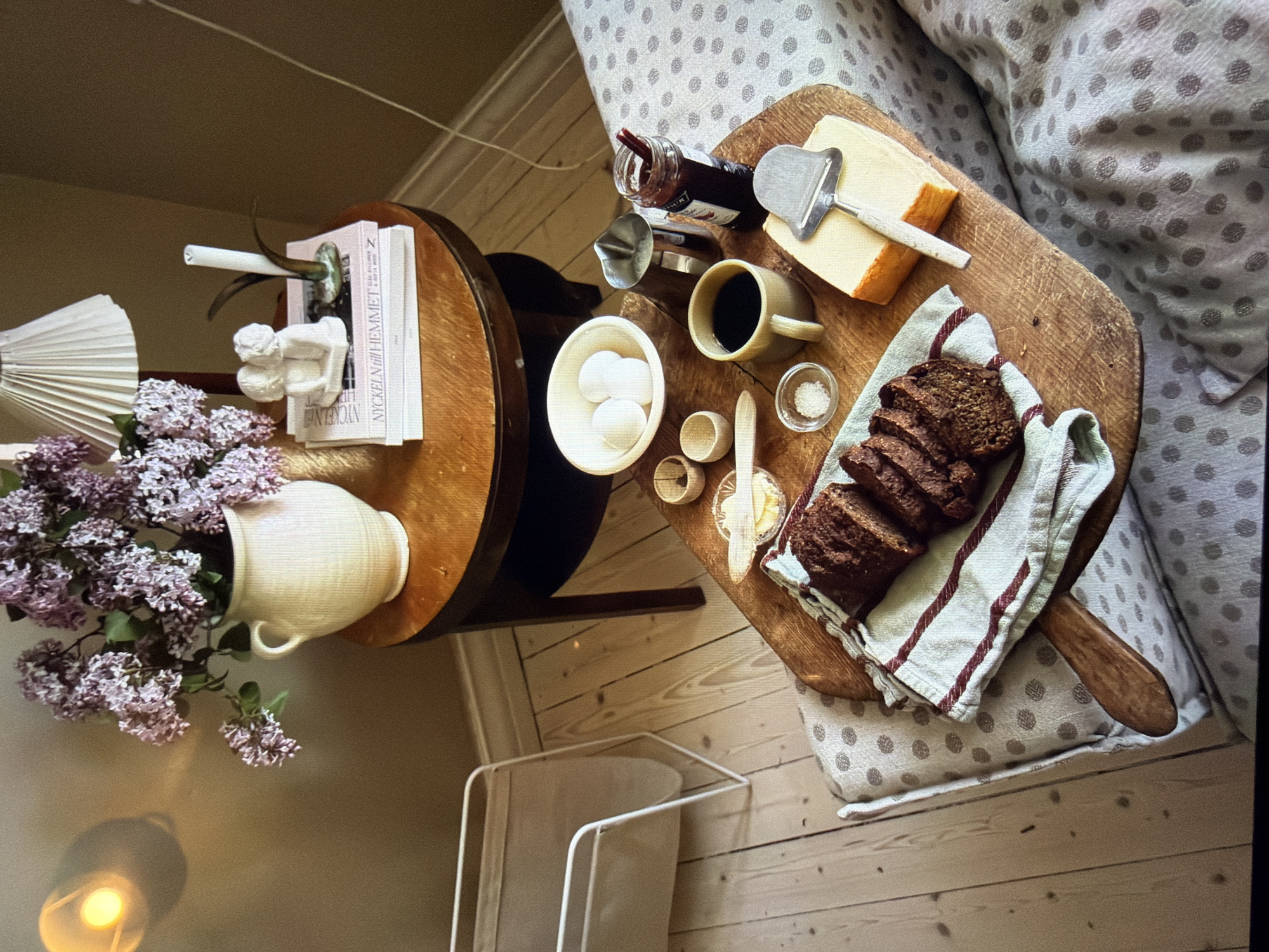 This indoor still life depicts a cozy breakfast or snack setup within a warm, inviting setting. The scene is captured from an overhead perspective, looking down onto a light wooden floor.
On the right side of the frame, a sofa or armchair with light-colored, polka-dotted upholstery holds a paddle-shaped wooden serving board. On this board, a meal is arranged: slices of dark bread rest on a striped linen towel, alongside a block of yellow cheese with a metal cheese slicer. A small jar of red jam with a spoon is visible next to the cheese. A light-colored mug contains coffee, and a small white bowl holds three white eggs. Complementing these are a tiny bowl of white salt, a small container with butter and a wooden spreading knife, and a small metal pitcher, possibly for milk or cream.
To the left, a round wooden side table holds a light-colored ceramic pitcher brimming with vibrant purple lilac flowers. A white, pleated lampshade stands nearby. Also on the table, a stack of books acts as a base for a small white ceramic figurine, appearing to be a cherub-like figure, which holds a slender white candle.
At the bottom of the frame, on the wooden floorboards, a light-colored metal rack, possibly for magazines or blankets, is visible. The lighting suggests daytime, contributing to the serene and comfortable atmosphere.
The visible text on one of the white book spines reads "NYCKELN till HEMMET," which is Swedish for "The Key to the Home."