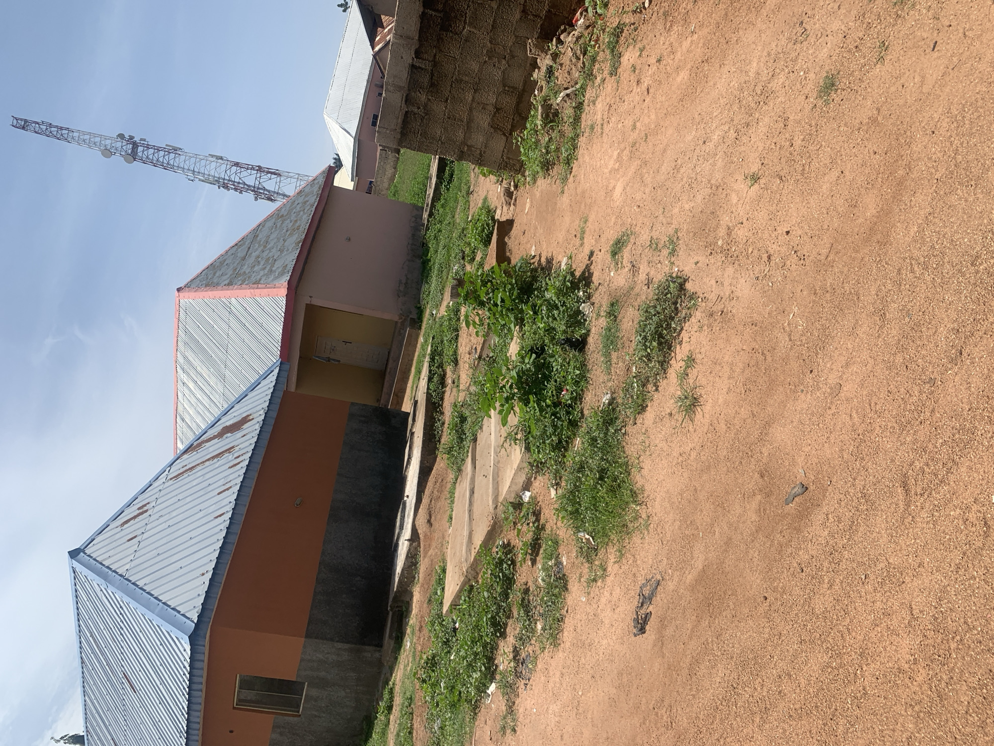 In Jauro Yinu, Nigeria, this image captures a scene on a sunny day. In the foreground, a dusty, reddish-brown dirt path leads into the frame, with sparse patches of green grass and scattered debris. To the left, a building with an orange and gray facade features a corrugated metal roof and a small window. Adjacent to it, another building with a pinkish-orange facade and a pink-trimmed corrugated metal roof is visible. In the background, a tall, latticed telecommunications tower with multiple antennas stands against a clear blue sky. Further back, other buildings with corrugated roofs are present, along with a brick wall in the mid-ground. The environment appears to be an outdoor setting with residential structures and infrastructure.