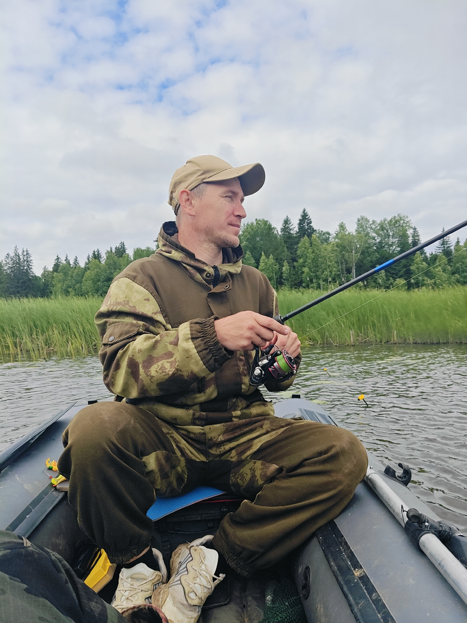 A man is fishing from an inflatable boat on a lake. He is wearing a camouflage jacket and a beige baseball cap. He is holding a fishing rod and reel. The boat is in a body of water with reeds and trees on the shore. The sky is cloudy. The scene appears to be in Detskii Dom (Bashenino), Russia.