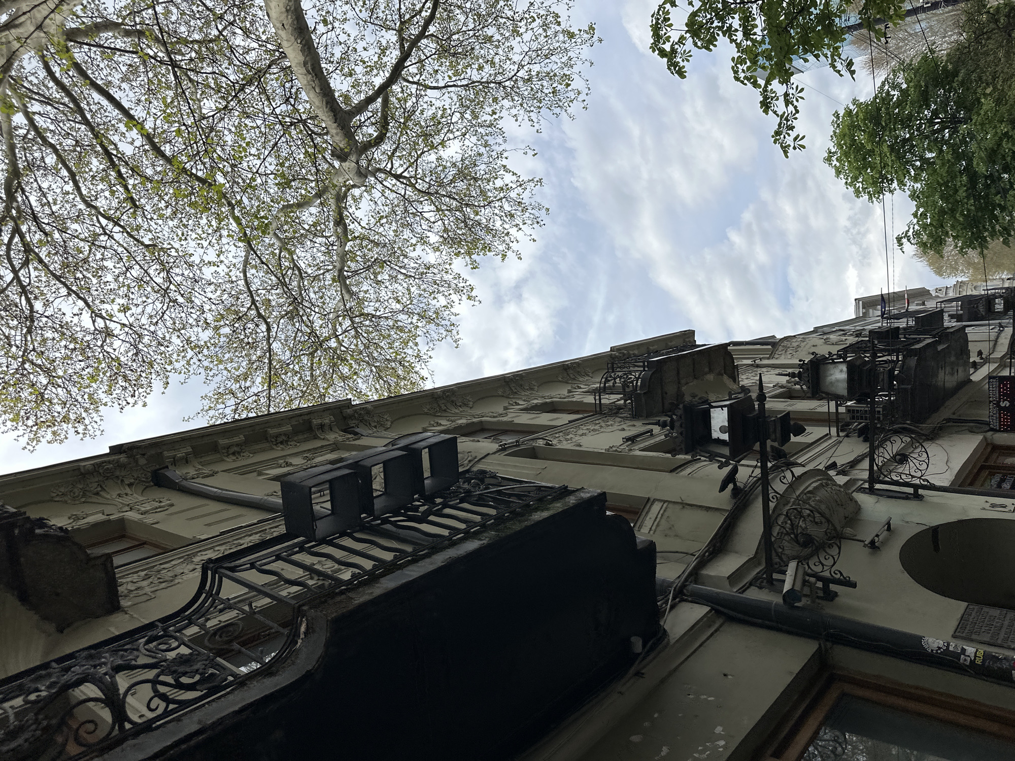 Looking up from a low angle, the photo shows a multi-story building facade in Tbilisi, Georgia, under a partly cloudy sky with trees visible above. The building is light-colored, likely stucco, with detailed architectural ornamentation along the roofline and around windows. Several black iron balconies with ornate railings are visible. Various air conditioning units and other mechanical fixtures are attached to the building's exterior. Wiring and pipes run along the facade. Stickers and small plaques are attached to the walls near the bottom right. The trees above have foliage in spring, showing new growth.