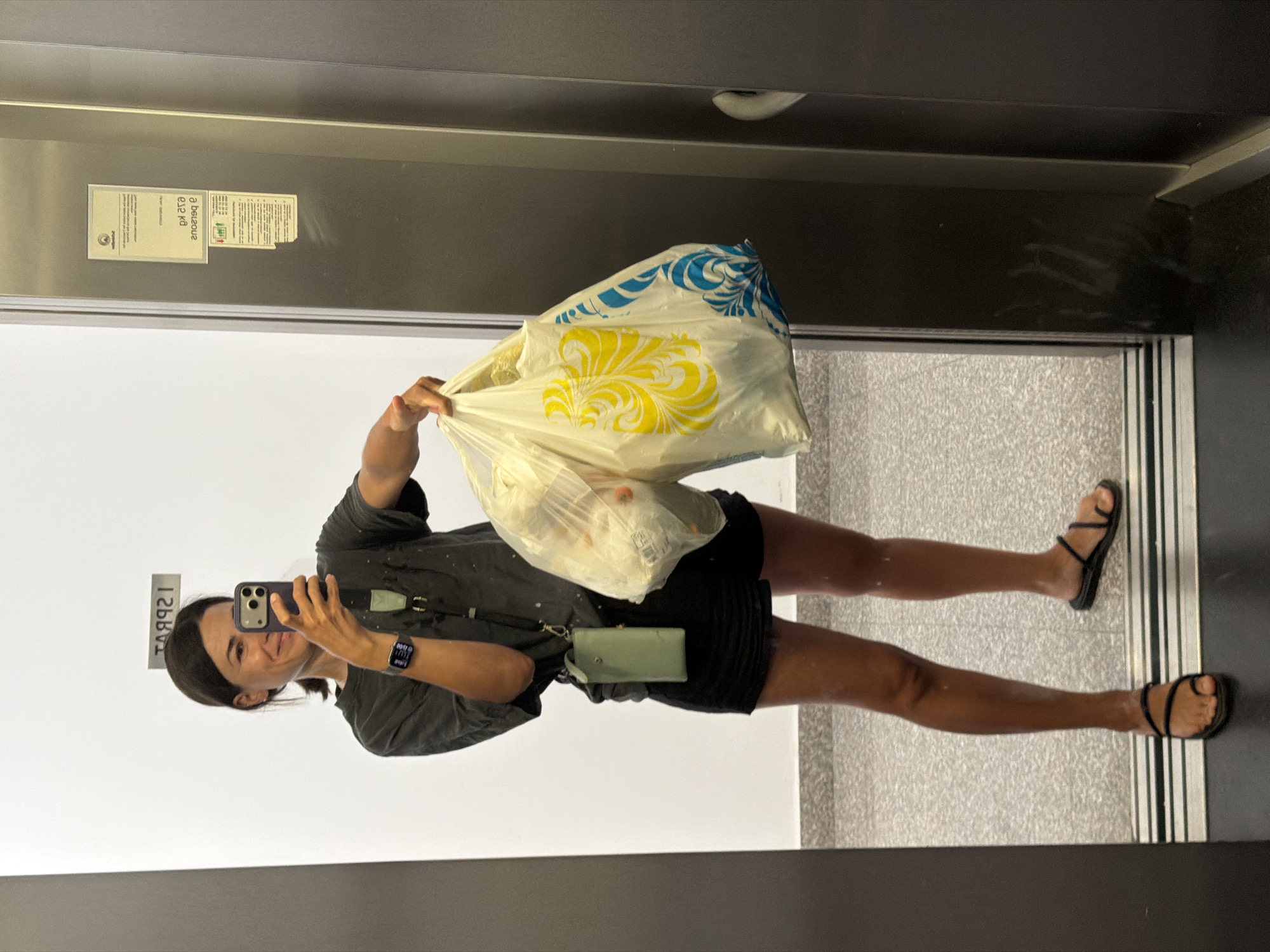 The image shows a woman taking a mirror selfie in an elevator. She is reflected full-body, holding a smartphone in her right hand, positioned to capture her reflection. In her left hand, she carries two plastic shopping bags; one large, light-colored bag features prominent yellow and blue swirl patterns, while a smaller, transparent bag holds what appears to be produce.
The woman is dressed in a dark t-shirt or loose top, black shorts, and black flat sandals with multiple straps. A light green or mint-colored cross-body purse hangs around her neck/shoulder. On her left wrist, she wears a smartwatch (appearing to be an Apple Watch) which clearly displays the time "14:00" and "PM", indicating 2:00 PM. She has dark hair, tied back, and a pleasant, slight smile.
The setting is an elevator, characterized by its metallic walls and a light-colored, speckled floor. Two rectangular signs are visible reflected on the elevator wall. The left sign features a logo on its left side and text that includes "аВ9аоus" (likely a mirrored Cyrillic word) and "еg ка". The right sign also contains some mirrored text, including "а d s 9", and features red and green graphical elements. Another small, thin rectangular plate is visible on the wall behind her head, reflecting the number "129842". Given the provided context, the location is Budva, Montenegro. The clothing suggests warm weather.