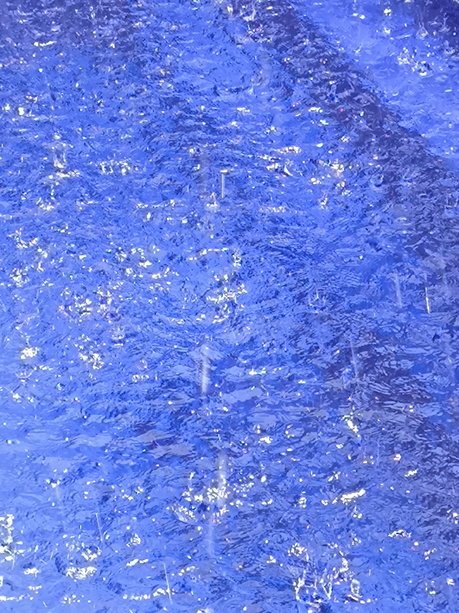 The image shows a close-up view of a body of water, possibly a pool or fountain, with a predominantly blue hue. The water's surface is rippled and reflects light in scattered, bright glints. Raindrops are falling and visible as vertical streaks or small splashes in the water. A darker, less reflective area is seen towards the top right, which may be the edge or bottom of the water feature. The location of the scene is Unknown, Unknown.