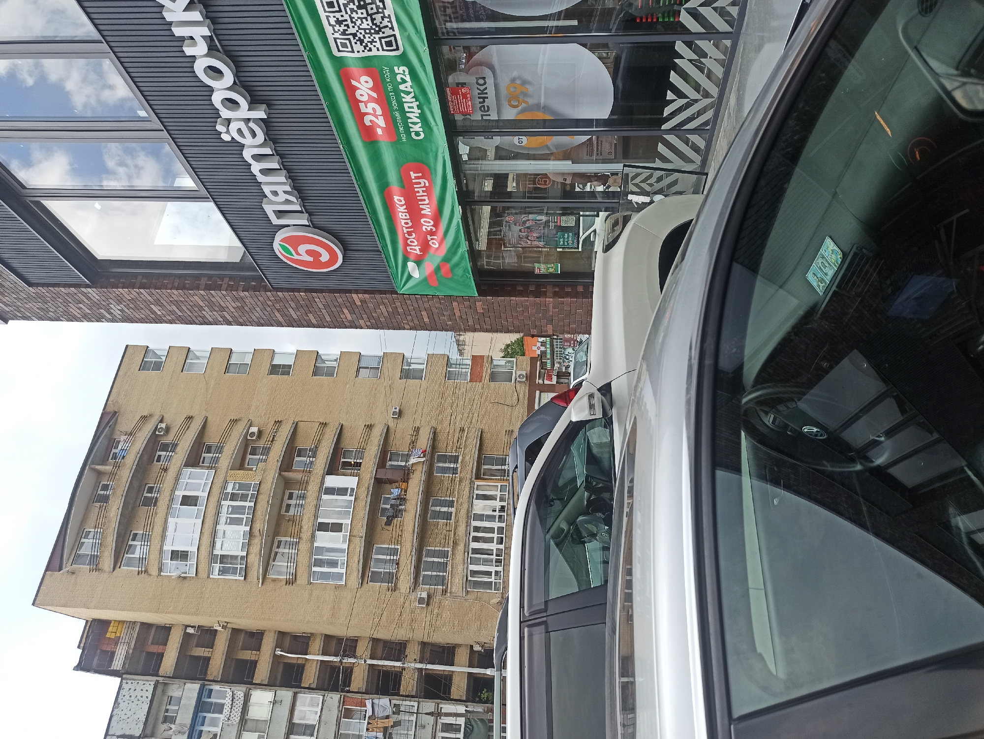 The image shows a street scene in Atabe, Georgia, with a focus on a "Pyaterochka" grocery store. The store has a prominent sign with the "Pyaterochka" logo, a red circle with a white "5" inside. A green banner is displayed, advertising "Delivery from 30 minutes" and a "-25% discount for the first order with code SKIDKA25". The store entrance has large windows, with some of the interior visible, including shelves with products and possibly some advertising posters.
In front of the store, several cars are parked. The perspective is from inside one of these cars, looking out. The interior of the car is visible, including the dashboard, steering wheel, and part of the passenger side. A sticker or decal is visible on the windshield.
Behind the store, a tall, multi-story building with a distinctive architectural style is visible. The building is made of light-colored brick or concrete and features a curved, cantilevered section with many windows. Laundry is hanging from some of the balconies on this building. The sky is overcast, suggesting a cloudy day. The overall impression is an urban street view, likely during daylight hours.