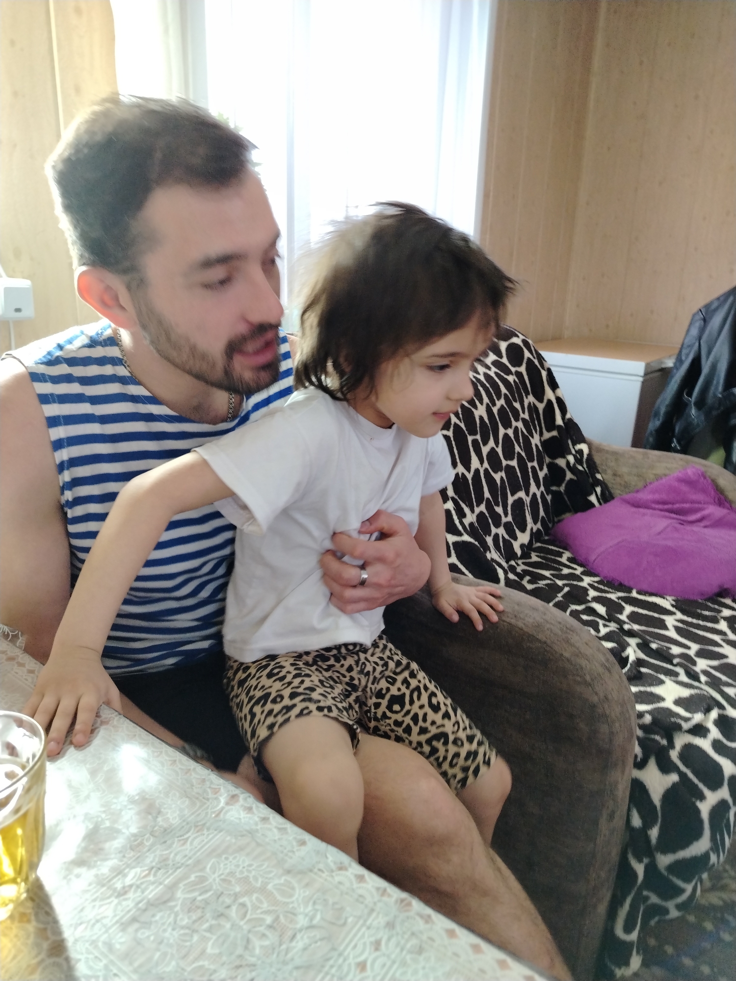 A man and a child are indoors in a room with a table, a sofa, and wooden walls. The man is seated and wearing a blue and white striped tank top and dark shorts. He has a beard. The child is sitting on the man's lap. The child wears a white t-shirt and leopard print shorts. The child's left hand rests on the table, and the man's left hand rests on the child's torso. The table has a light-colored patterned tablecloth, and a glass with a yellow liquid stands on it. Behind the man and child is a sofa with a brown patterned seat and a throw with a giraffe print. A purple pillow is on the sofa. A white cabinet is visible in the background. The jacket lies next to the cabinet. The image was taken in Большая Уса, Russia.