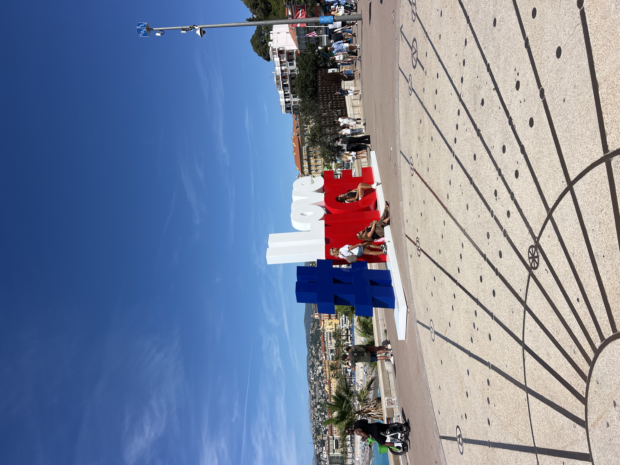 This image depicts the Promenade des Anglais in Nice, France. A large, three-dimensional sculpture spelling out "#LOVE" is centrally located, with the "#" symbol in blue and the word "LOVE" in white and red respectively. Several individuals are interacting with the sculpture and the surrounding area. On the left, a man is riding a green electric scooter. To the right of the sculpture, a woman is posing, and another woman is seated. In the background, a bustling promenade is visible with other pedestrians, buildings of Nice's coastline, palm trees, and the bright blue Mediterranean Sea under a clear sky. The foreground shows the patterned pavement of the promenade, casting distinct shadows from the sun.
