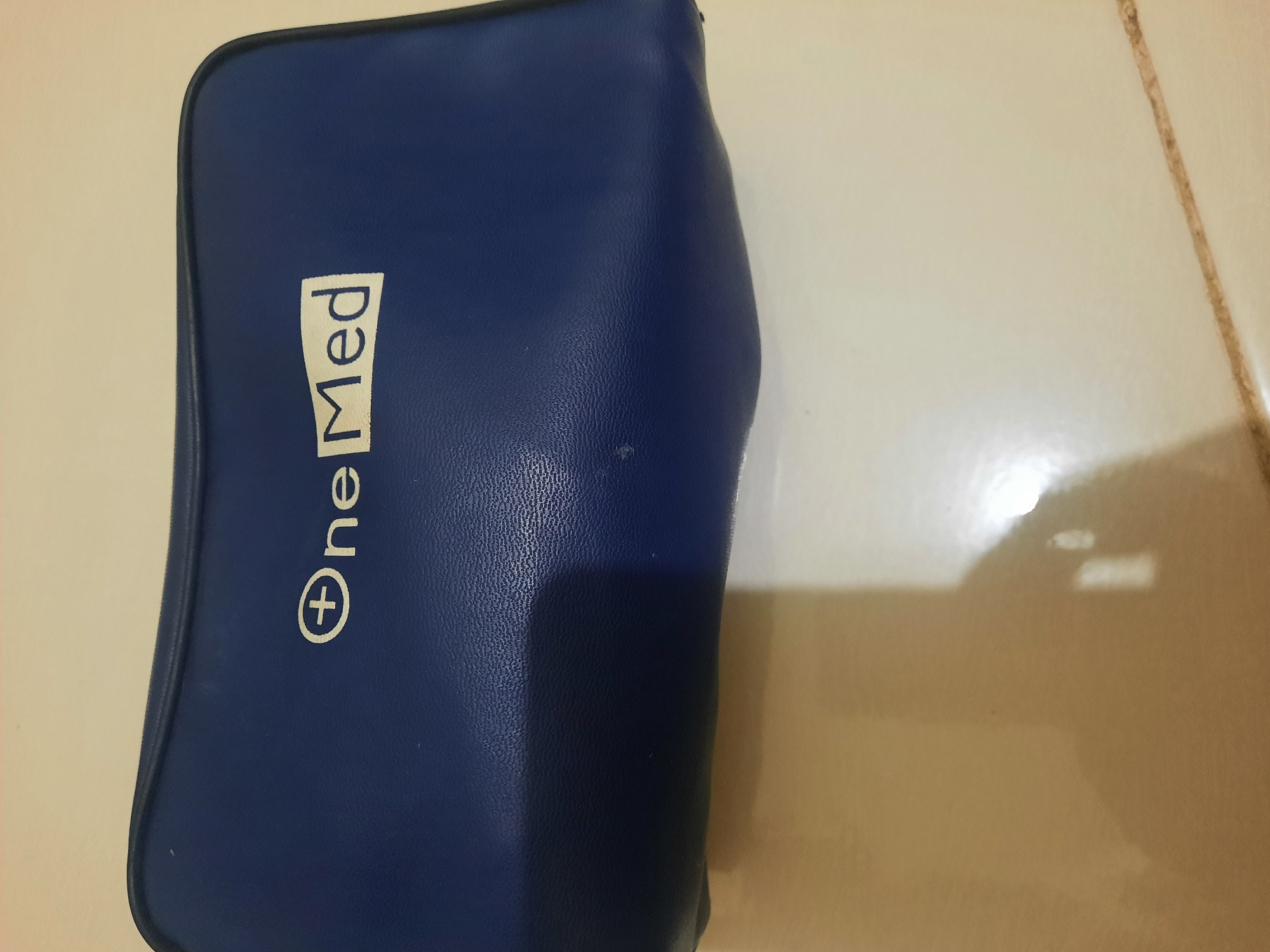 A dark blue pouch with a white logo is placed on a light-colored tiled surface. The logo consists of a white circle with a white cross inside it, followed by the text "ne Med" in white capital letters. The pouch appears to be made of synthetic leather and has a textured surface. The lighting in the image creates a shadow on the tiled surface, indicating the pouch is likely on a table or counter. The environment is an indoor setting, potentially a home or a commercial space, in Lhokseumawe, Indonesia. There are no people or animals visible in the image.