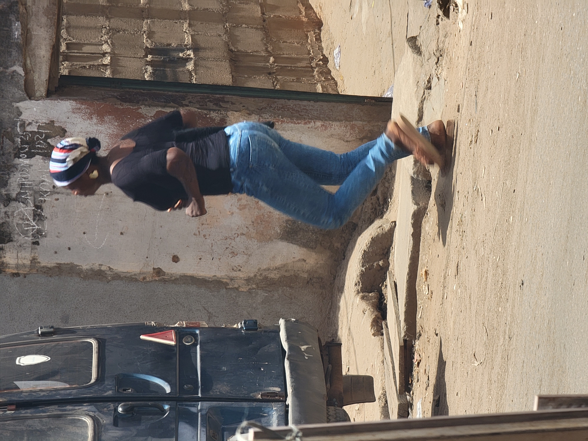 The image shows a woman standing on a concrete sidewalk next to a blue vehicle. The woman is wearing a black top, blue jeans, and brown shoes, and has a striped headwrap. She appears to be looking at something on the ground or interacting with the vehicle. The background includes a concrete wall with a textured top portion, suggesting a building or structure. The environment is dusty and urban, consistent with a location in Maiburiji, Nigeria.