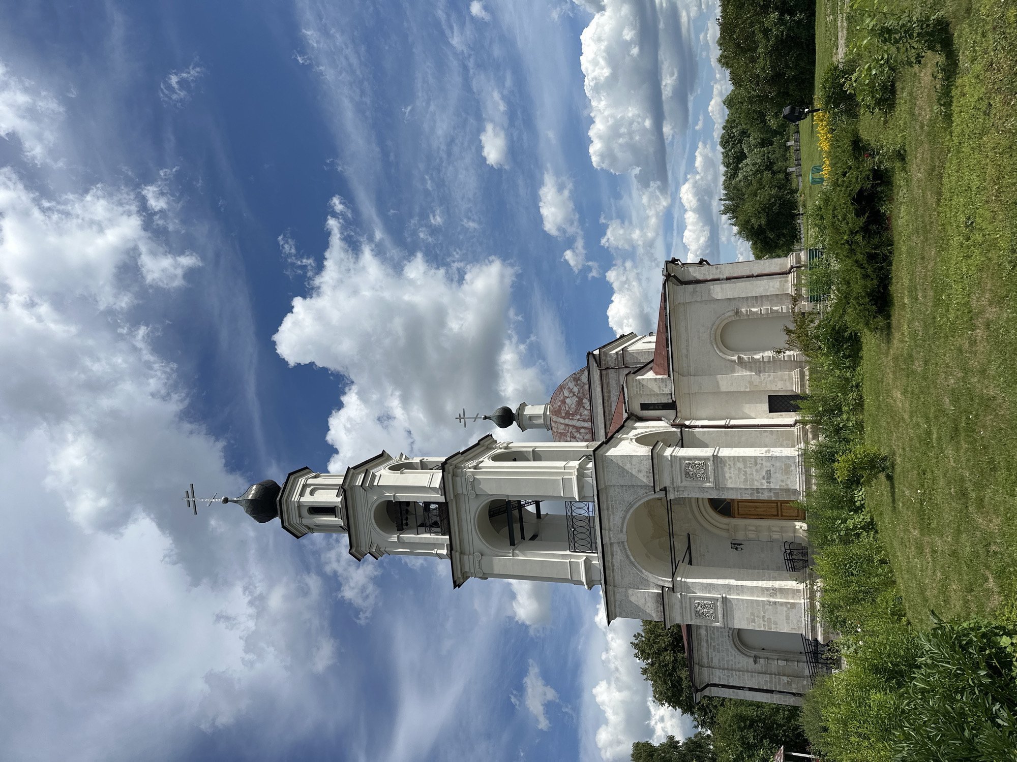 The image shows a white church with a bell tower against a bright blue sky with dramatic clouds. The church features arched windows and doorways, and the bell tower has multiple tiers of arches. A cross adorns the top of the bell tower and another dome. To the right of the church, there is a well-maintained grassy area with lush green bushes and trees. In the background, more trees and a hint of a fence are visible. The scene is illuminated by bright sunlight, suggesting it is daytime. The sky indicates it might be a partly cloudy day, with large, white clouds scattered across the blue. The overall impression is one of a peaceful and well-kept religious site in a rural or park-like setting. There is no visible text.